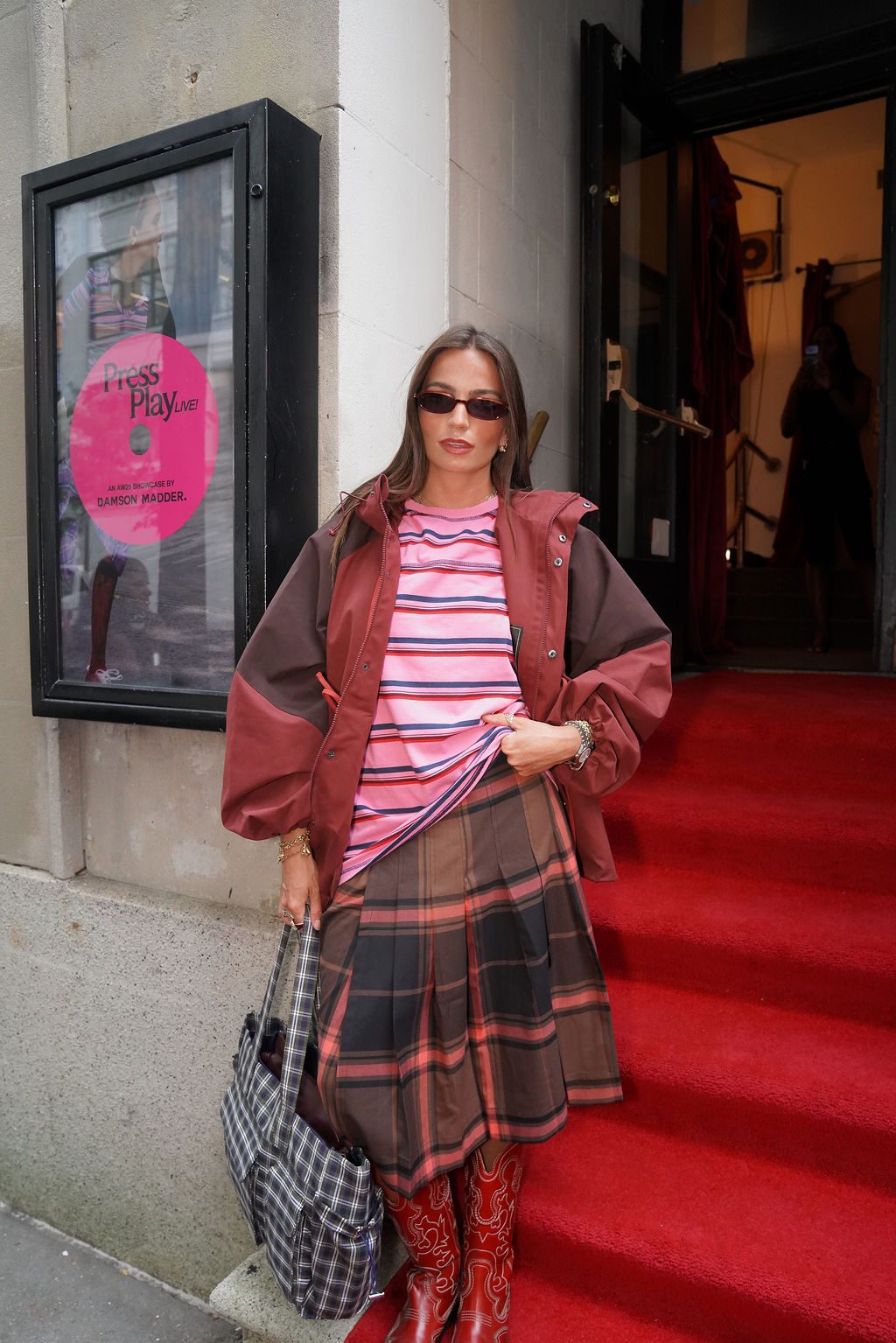 Pink striped tee, plaid skirt, red cowboy boots, and reddish-brown jacket on red carpet.