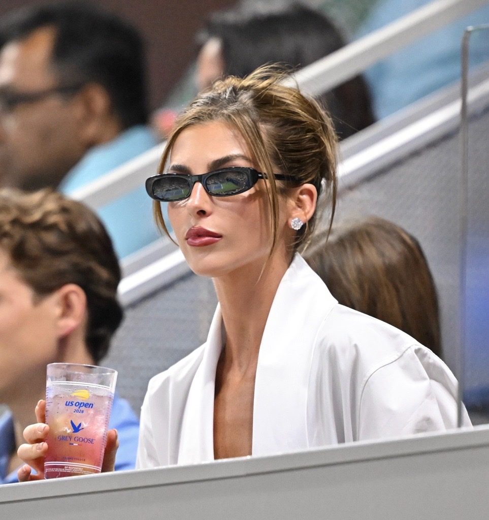 White draped top, dark sunglasses, and diamond earrings, holding a pink Grey Goose cocktail.