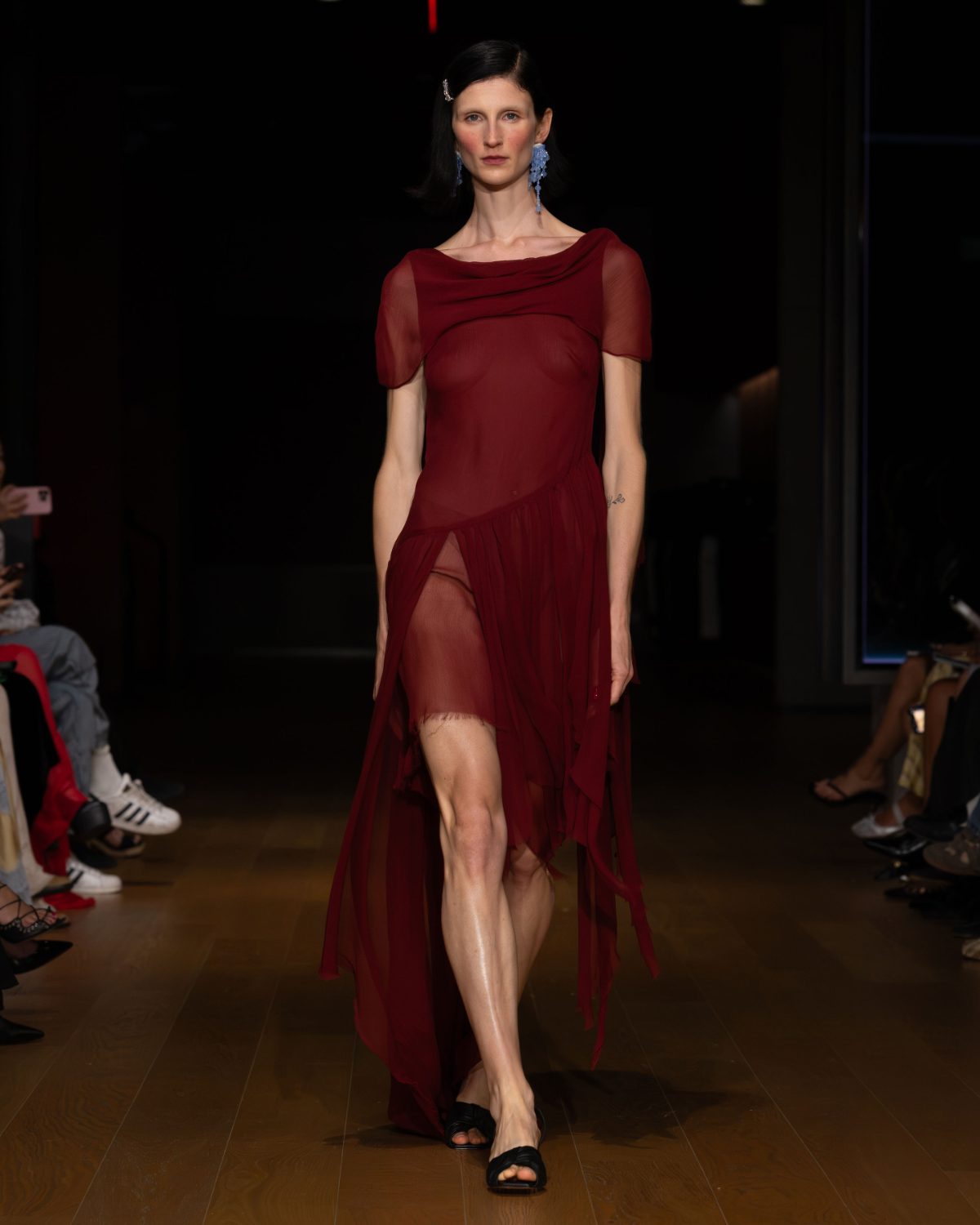 Sheer red asymmetric dress with blue dangling earrings and black flat sandals on a runway.