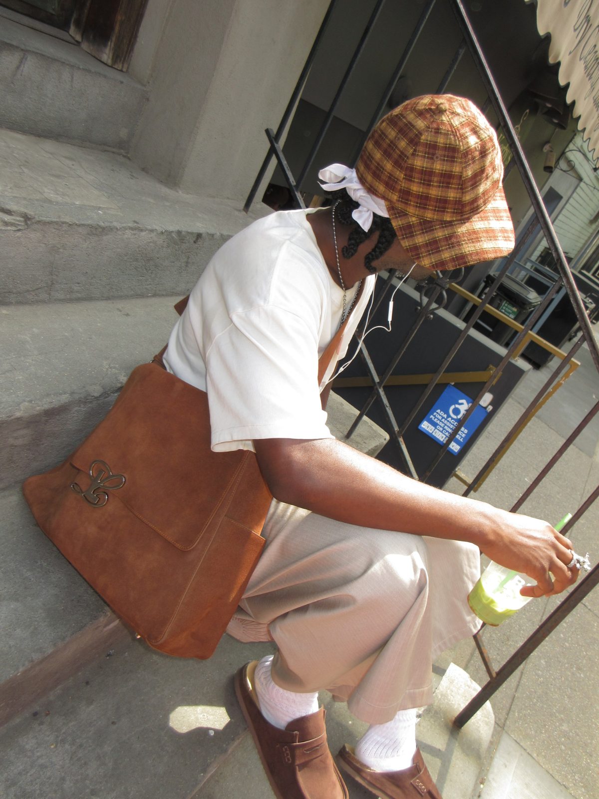 linging a large warm brown suede shoulder bag over a white tee, relaxed beige trousers, brown loafers, and a plaid cap
