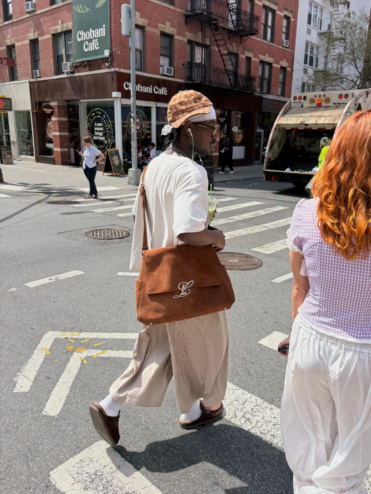 linging a large warm brown suede shoulder bag over a white tee, relaxed beige trousers, brown loafers, and a plaid cap