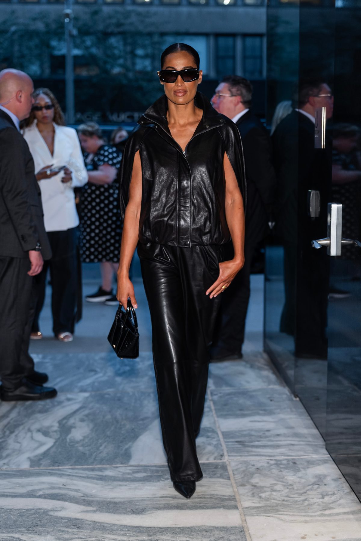 Black leather cape jacket and wide-leg trousers, pointed heels, and mini black handbag.