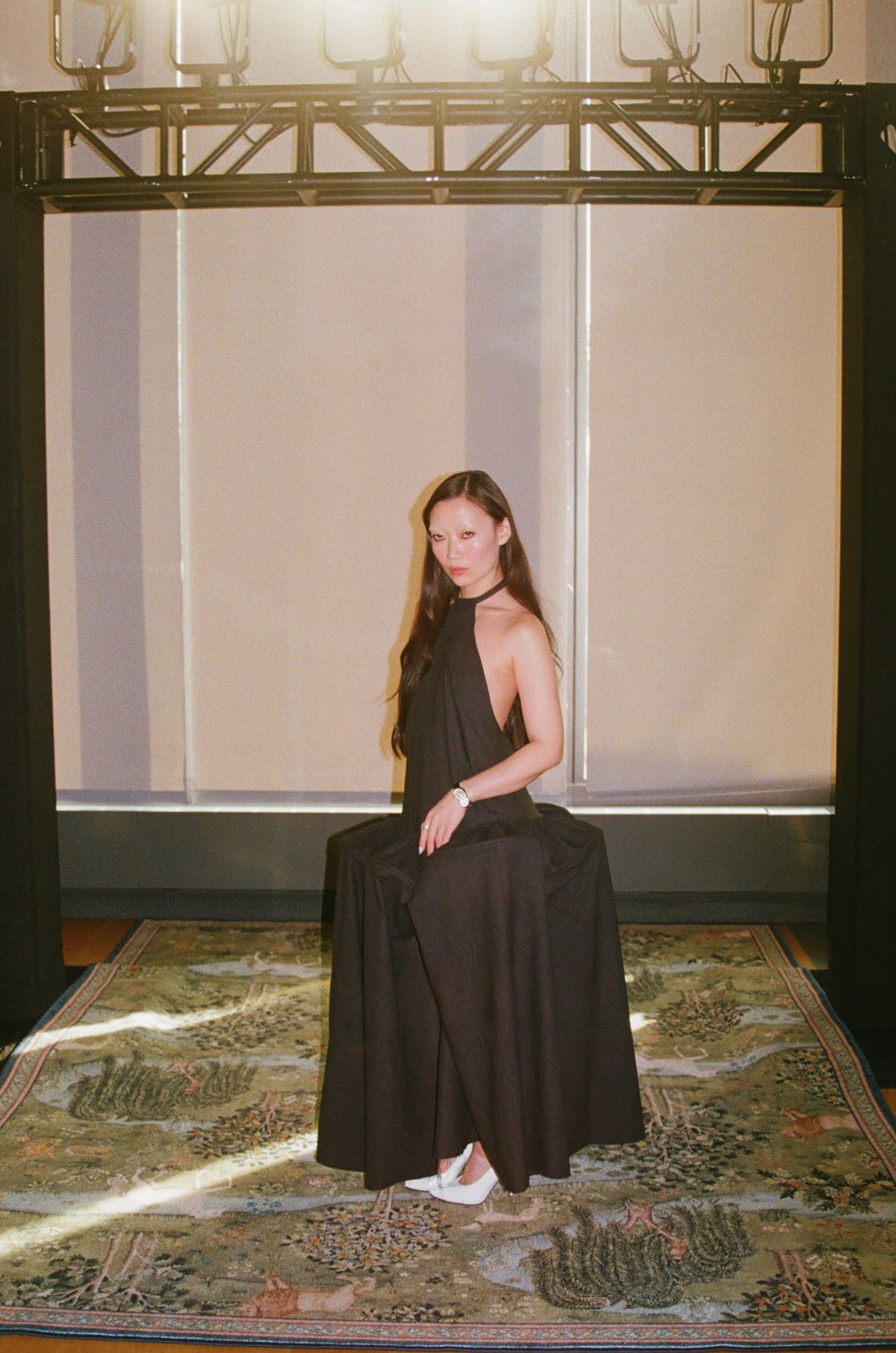 Black halter neck dress, white heels, seated on a patterned green carpet, fashion aesthetic.
