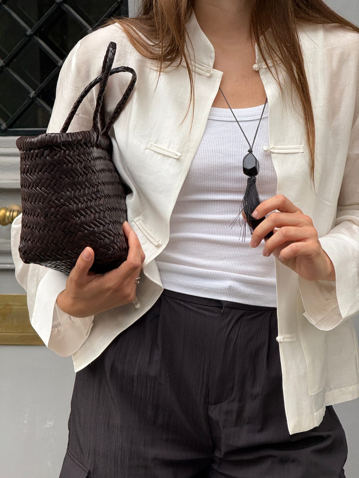 White ribbed top with an off-white silk jacket, black pants, a woven brown bag, and a black tassel necklace.