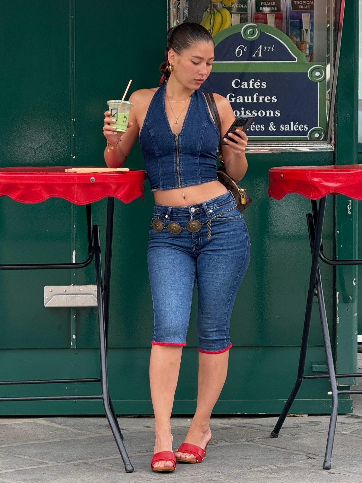 Denim halter top and cropped jeans with a western-style belt, holding a drink and phone.