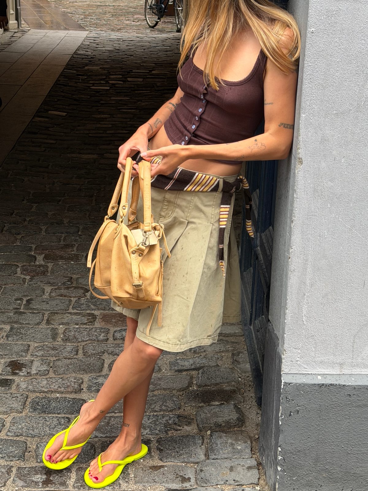Brown button-front tank top, khaki shorts, striped belt, yellow flip-flops, and tan handbag.