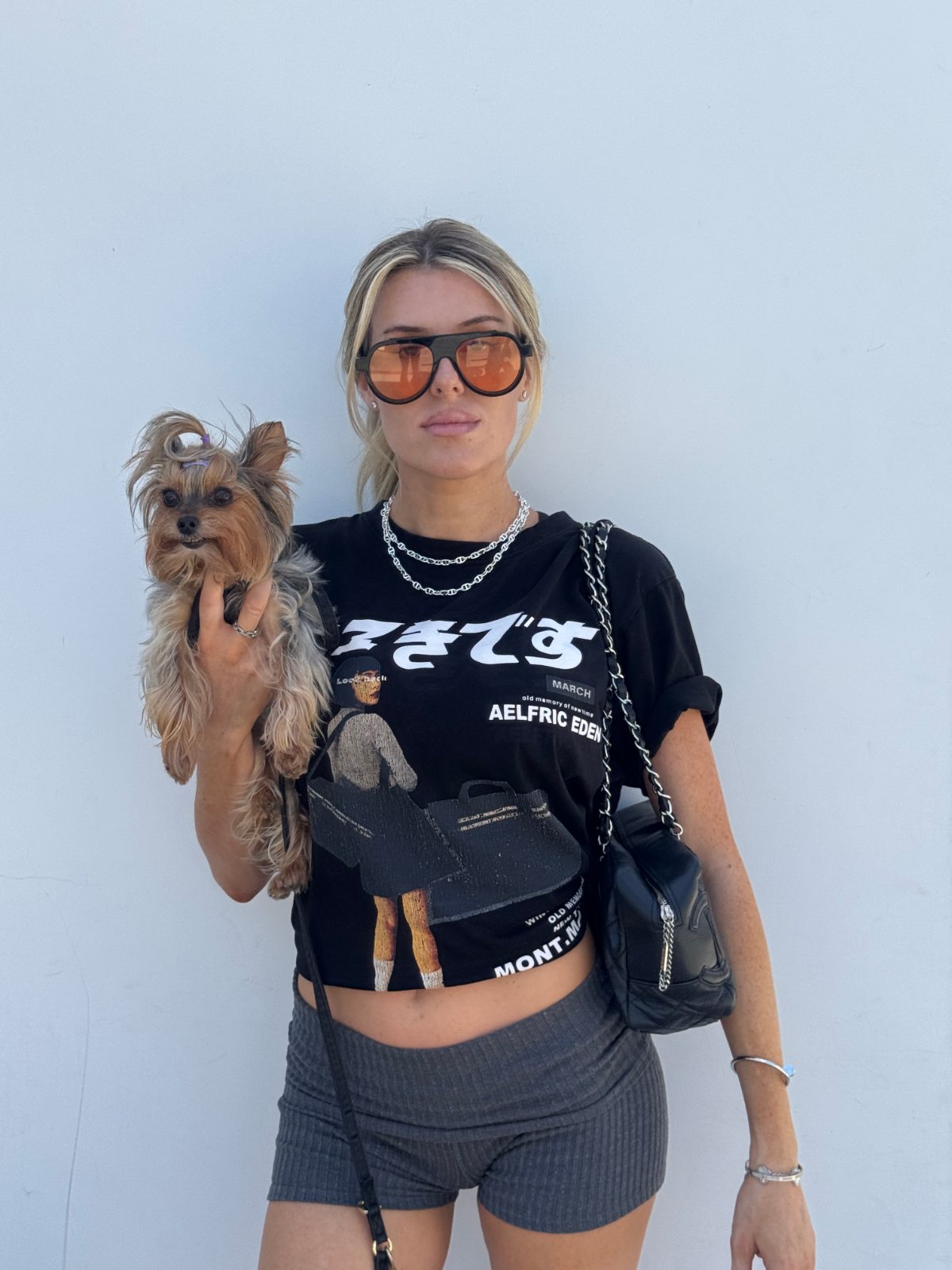 Black graphic crop top, grey ribbed shorts, and orange aviator sunglasses, holding a small dog.