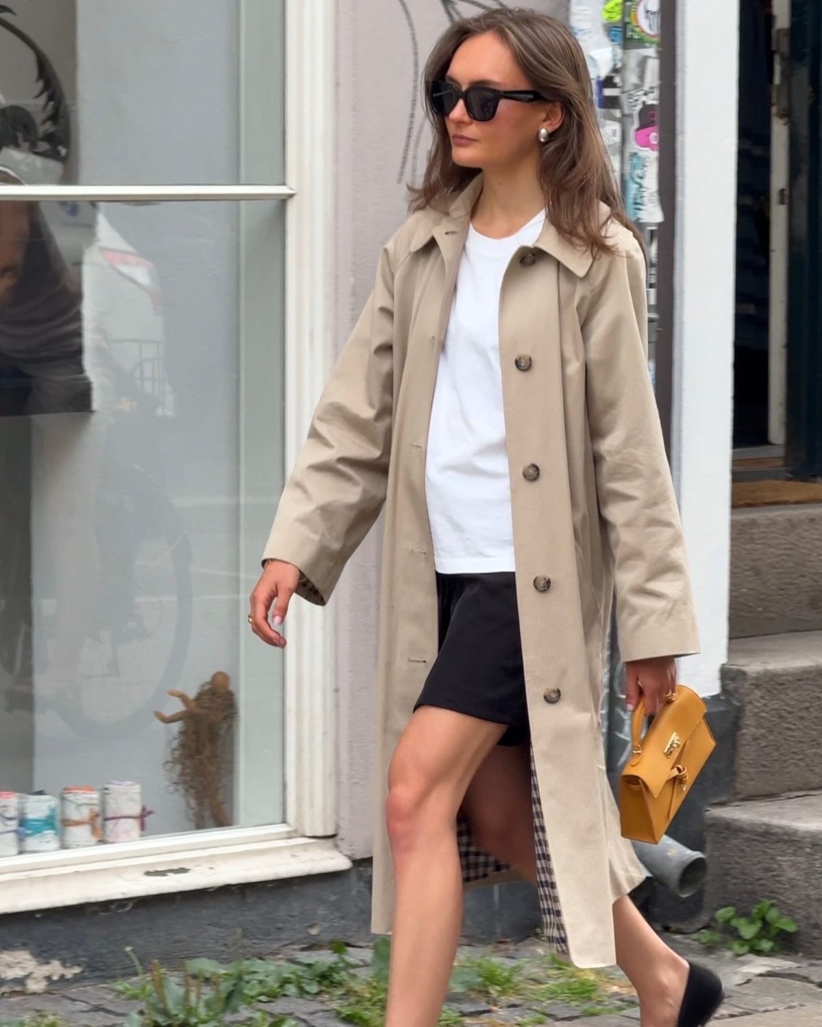 Beige trench coat with white t-shirt, black shorts, sunglasses, and a mustard yellow purse.