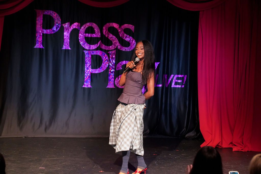 Smiling woman on stage in a gray peplum top, plaid midi skirt, and red heels.
