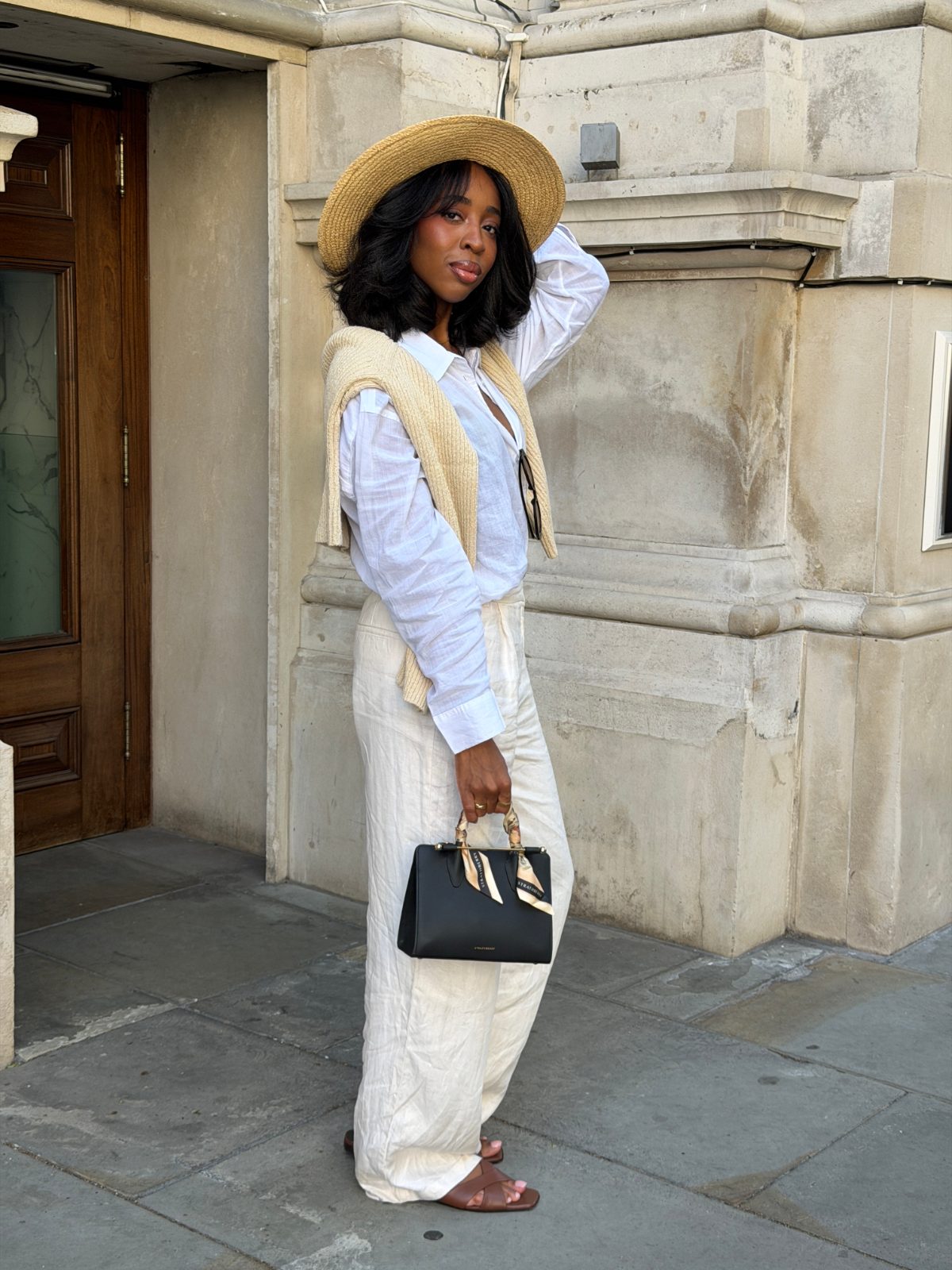 White button-up shirt, beige sweater, cream wide-leg pants, straw hat, and black handbag.