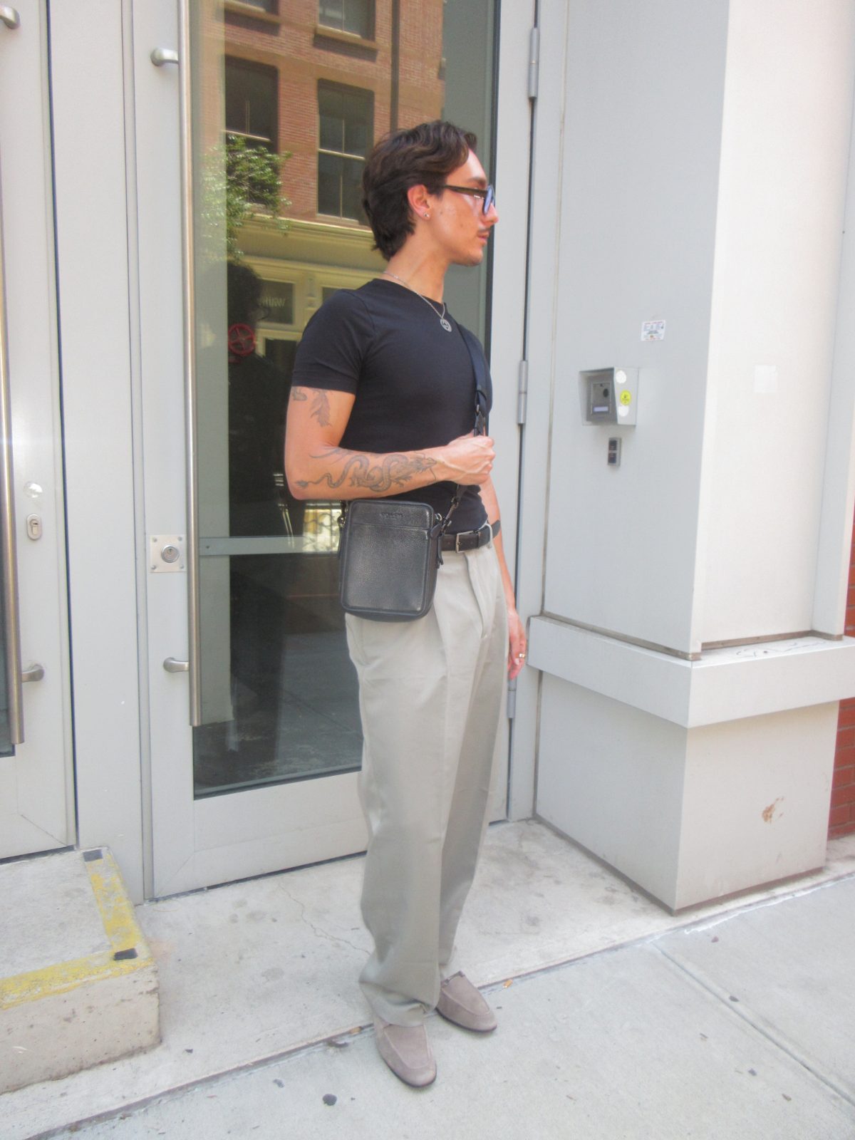 high-waisted beige pleated trousers with a fitted black crewneck, a black leather belt, grey suede loafers, and dark-rimmed glasses
