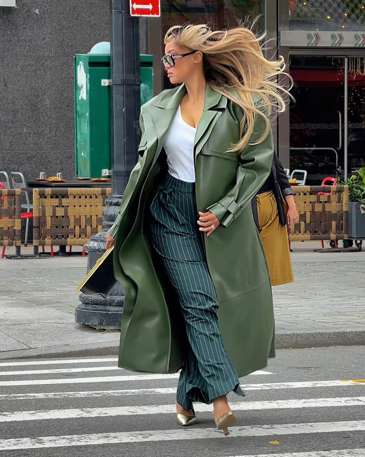 Green leather trench coat, white top, pinstripe pants, and gold heels on a city street.