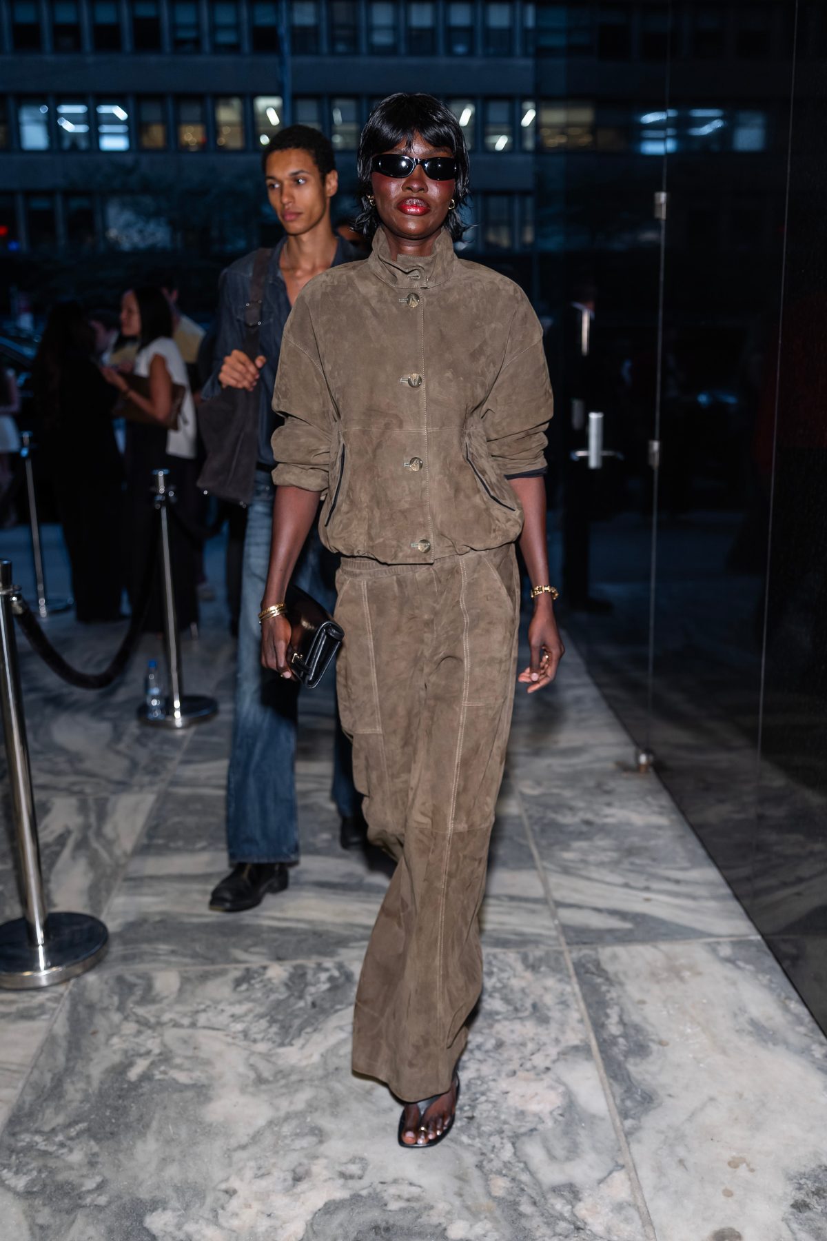 Brown suede jacket and wide-leg pants with black sunglasses and a matching clutch.