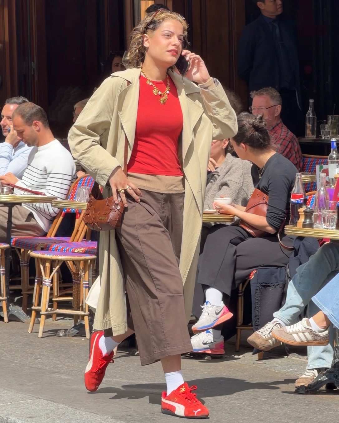 Khaki trench coat over a red top, brown trousers, and red sneakers at an outdoor cafe.