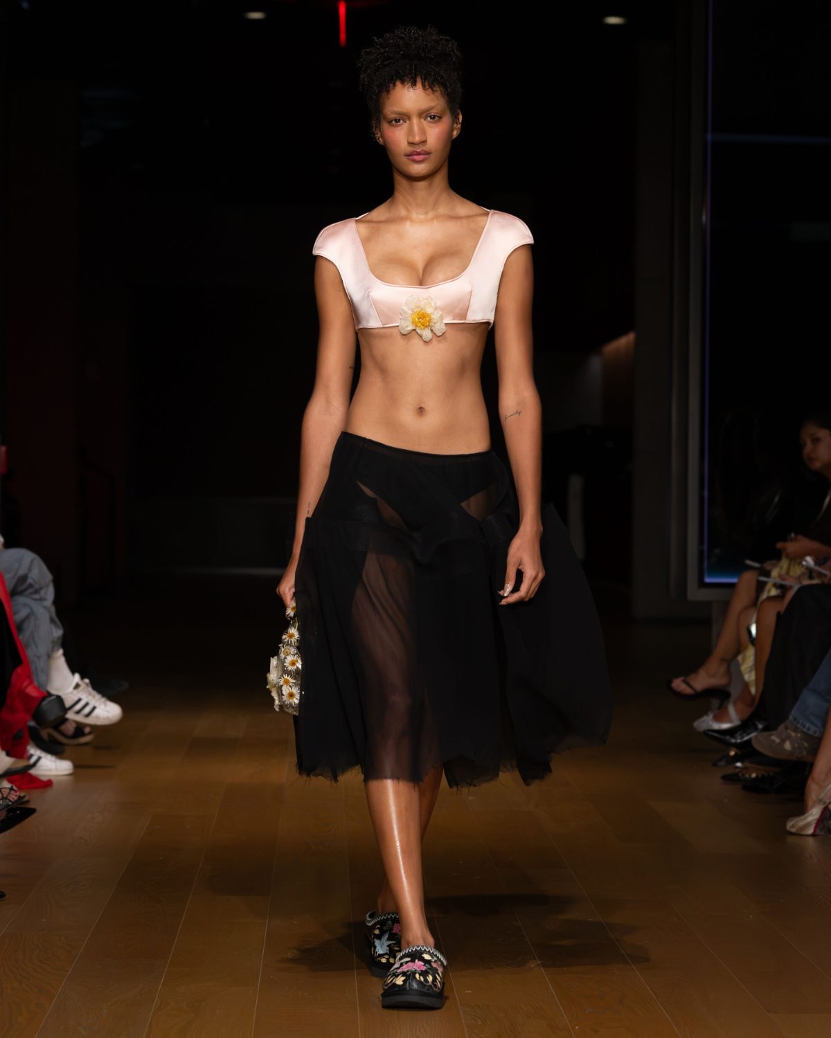 Pale pink crop top with a yellow flower appliqué, black sheer maxi skirt, and floral clogs.