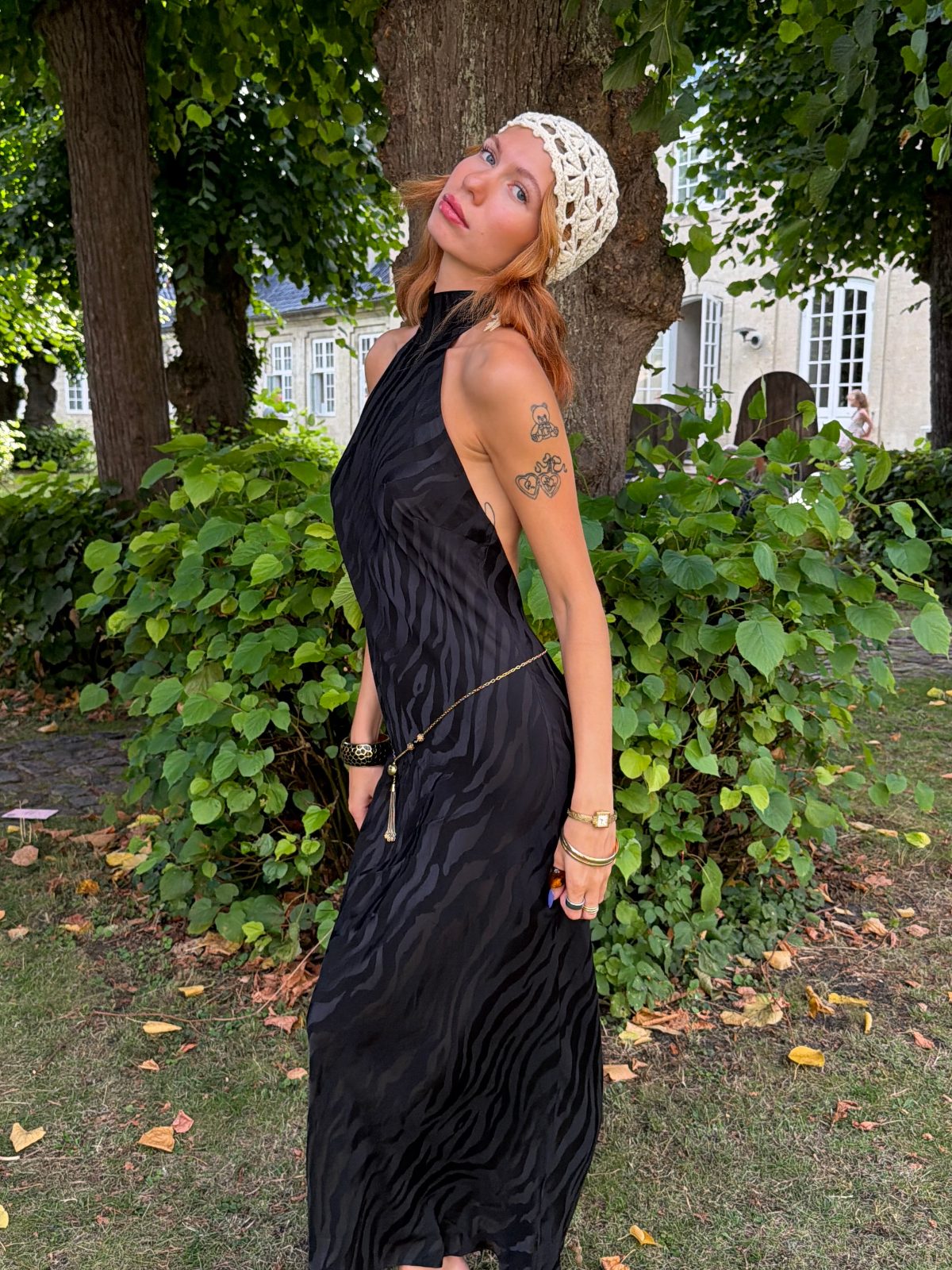 Black halterneck maxi dress with subtle zebra stripes, gold chain belt, and a white crochet hat.