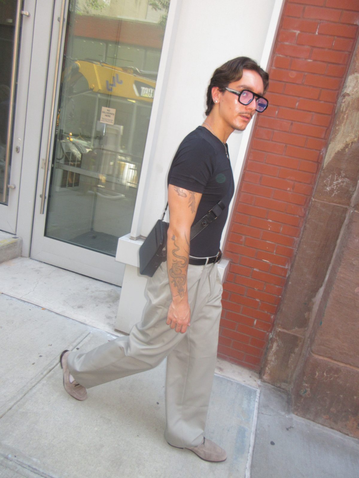 high-waisted beige pleated trousers with a fitted black crewneck, a black leather belt, grey suede loafers, and dark-rimmed glasses