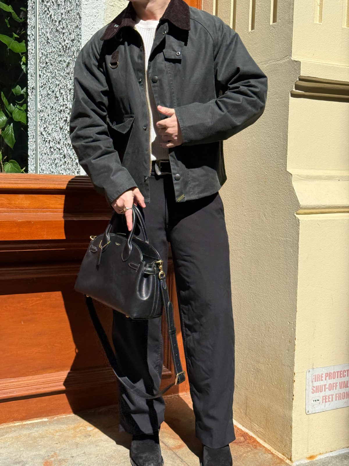 Dark green waxed cotton jacket with corduroy collar, white knit sweater, black pants, and handbag.