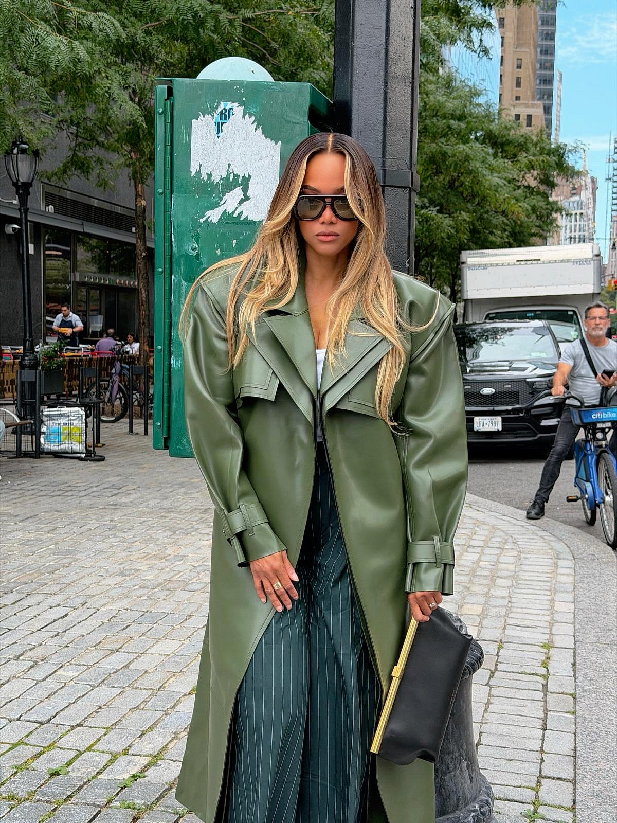 Olive green faux leather trench coat, pinstripe pants, and black clutch on city street.