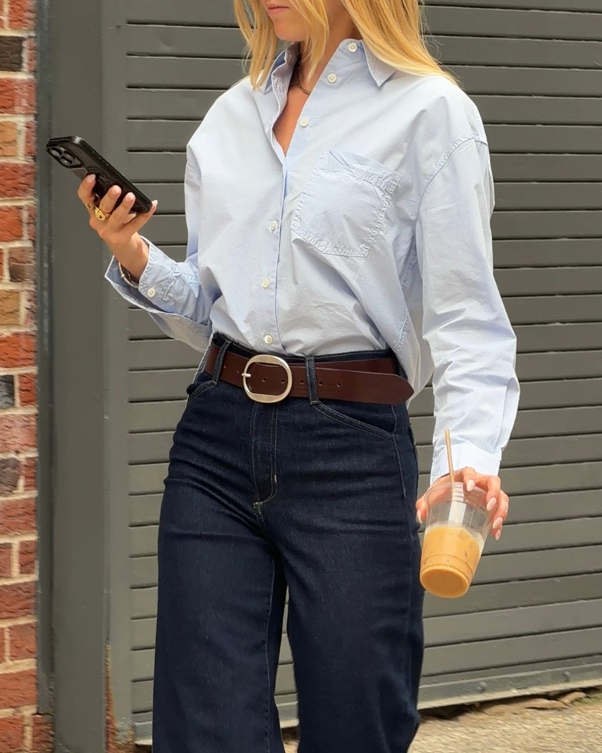 Light blue button-up shirt, dark wash jeans, brown belt, holding phone and iced coffee.