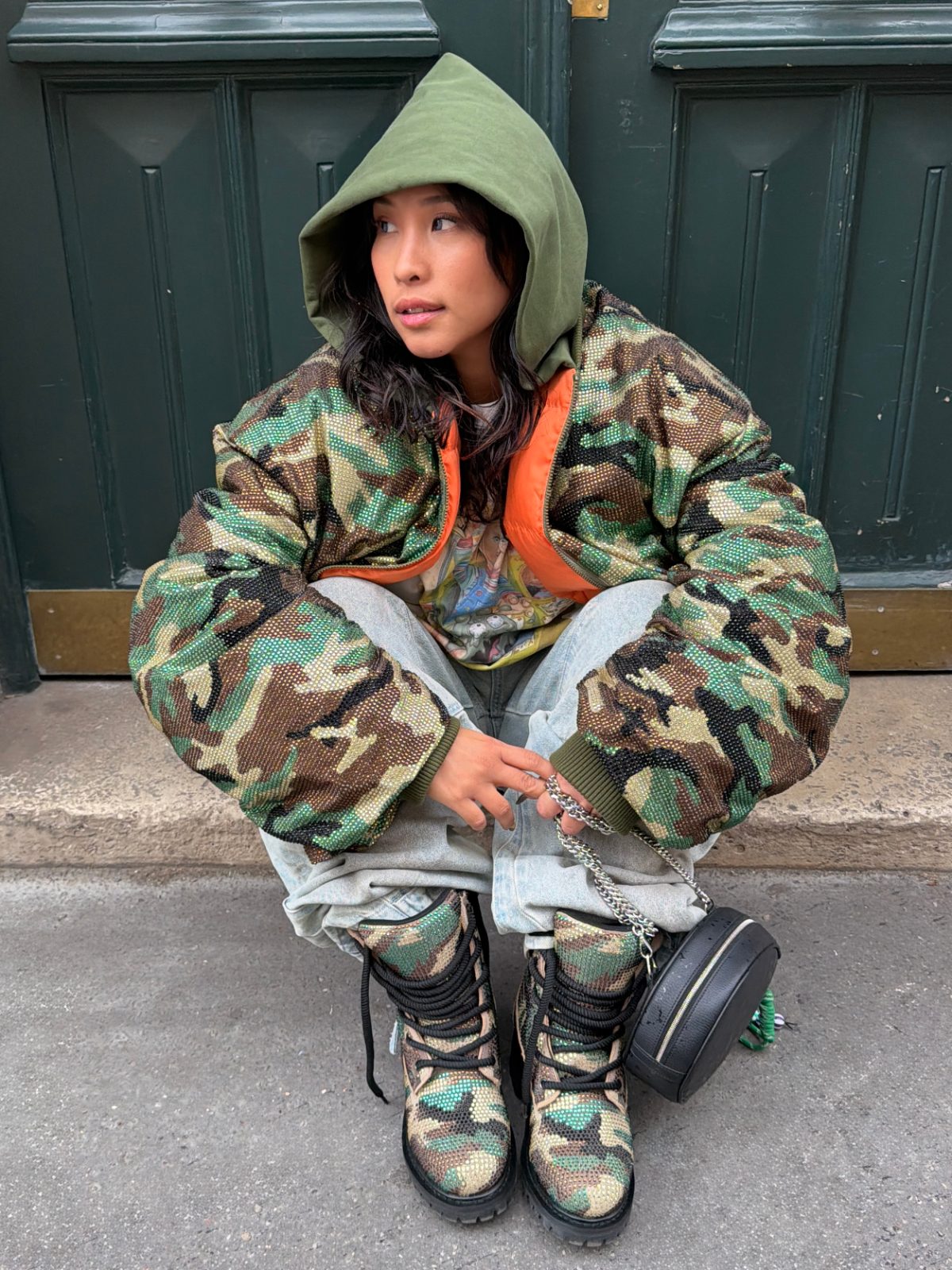 Camo puffer jacket with green hoodie, acid wash jeans, matching boots, and black handbag.