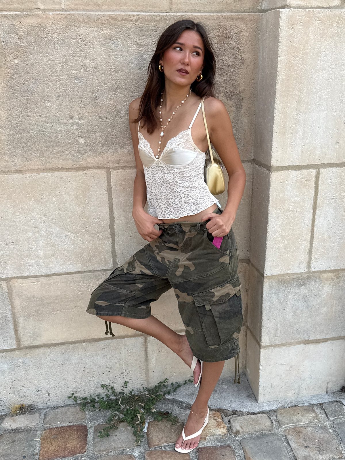 White lace corset top, camo cargo shorts, and white flip-flops, leaning against a stone wall.