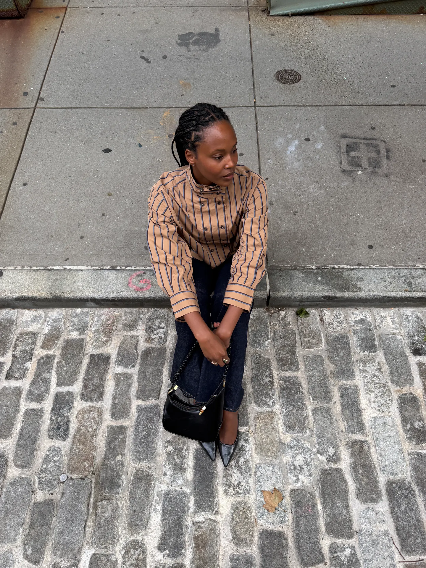 Tan striped button-up shirt, dark jeans, black purse, and pointed heels on cobblestones.