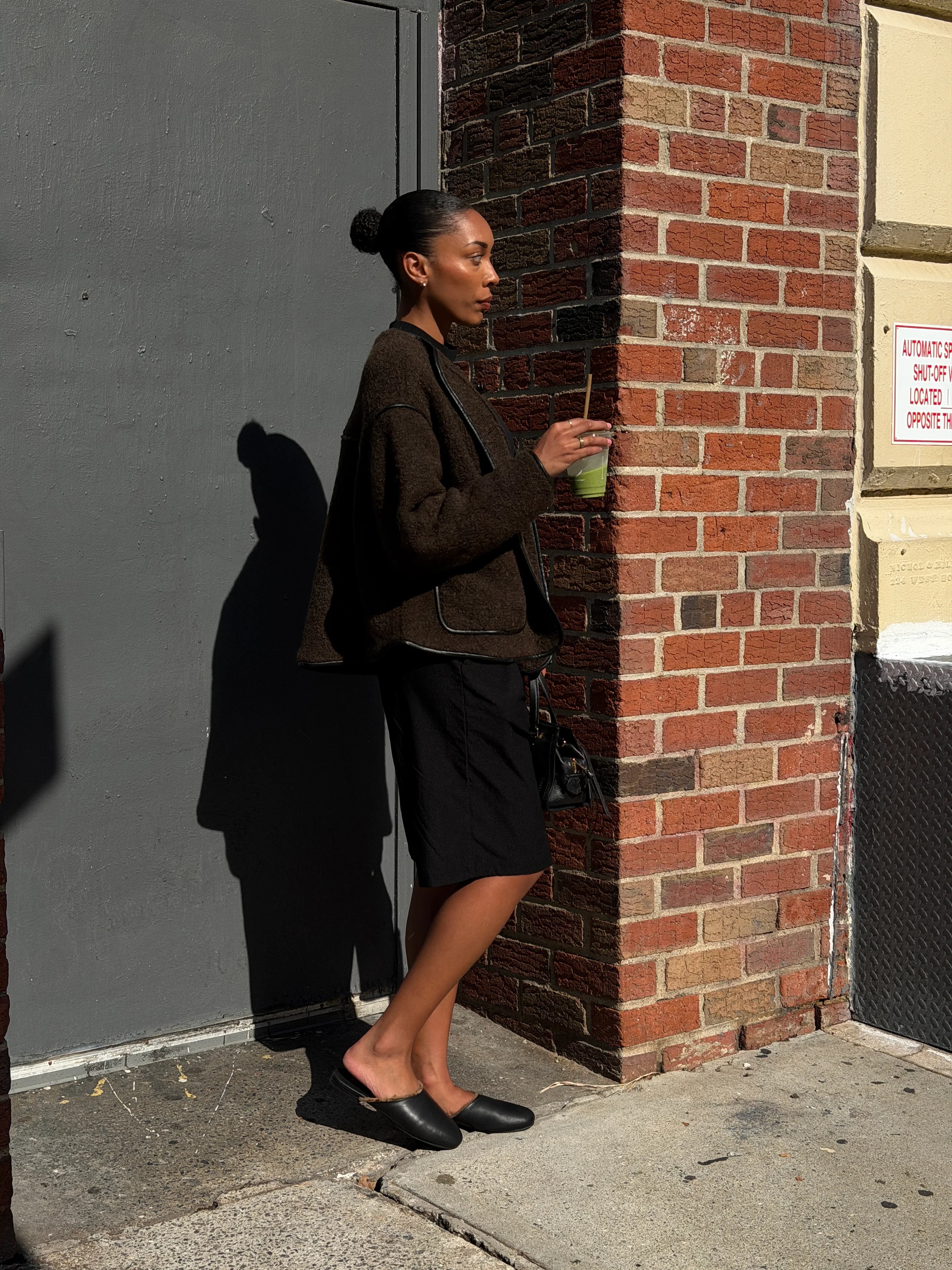 Brown textured jacket, black shorts, and black mules against a brick wall.