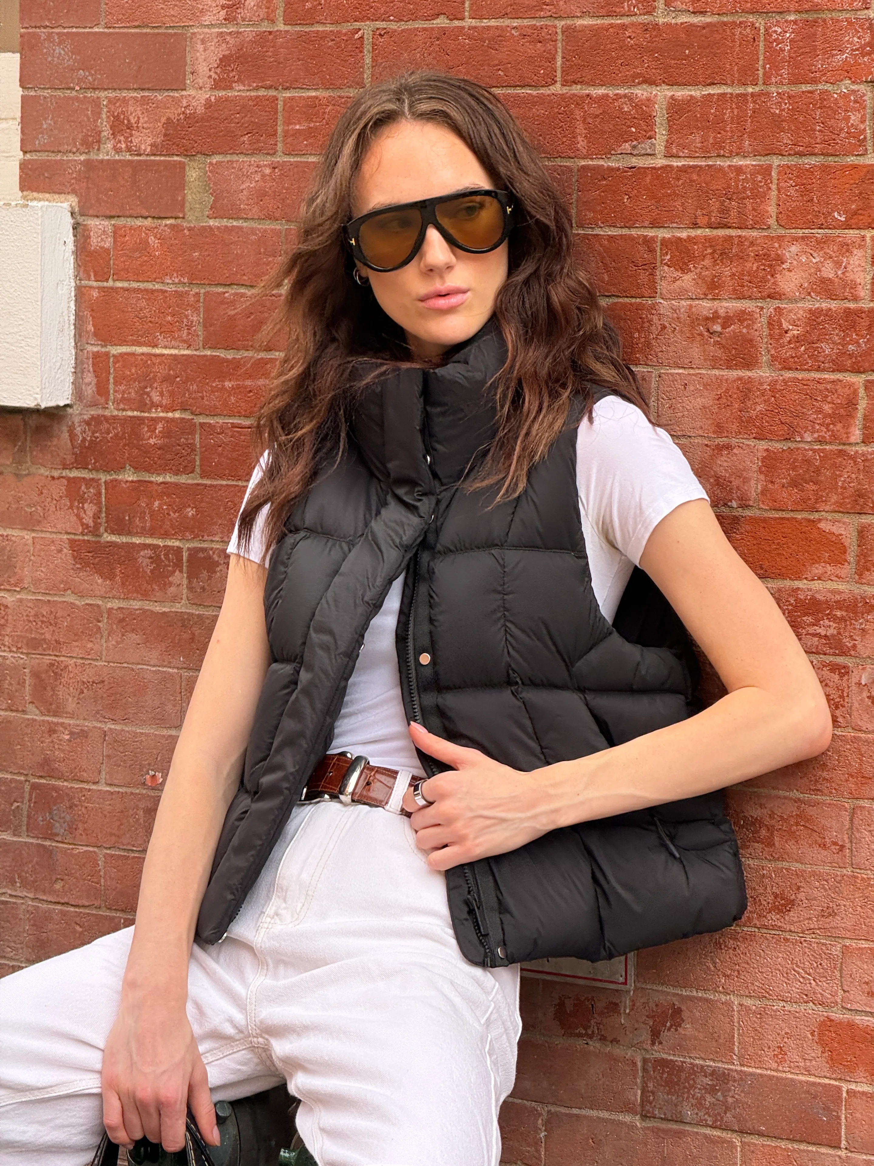 Black puffer vest, white tee, white jeans, brown belt, and aviator sunglasses against a brick wall.