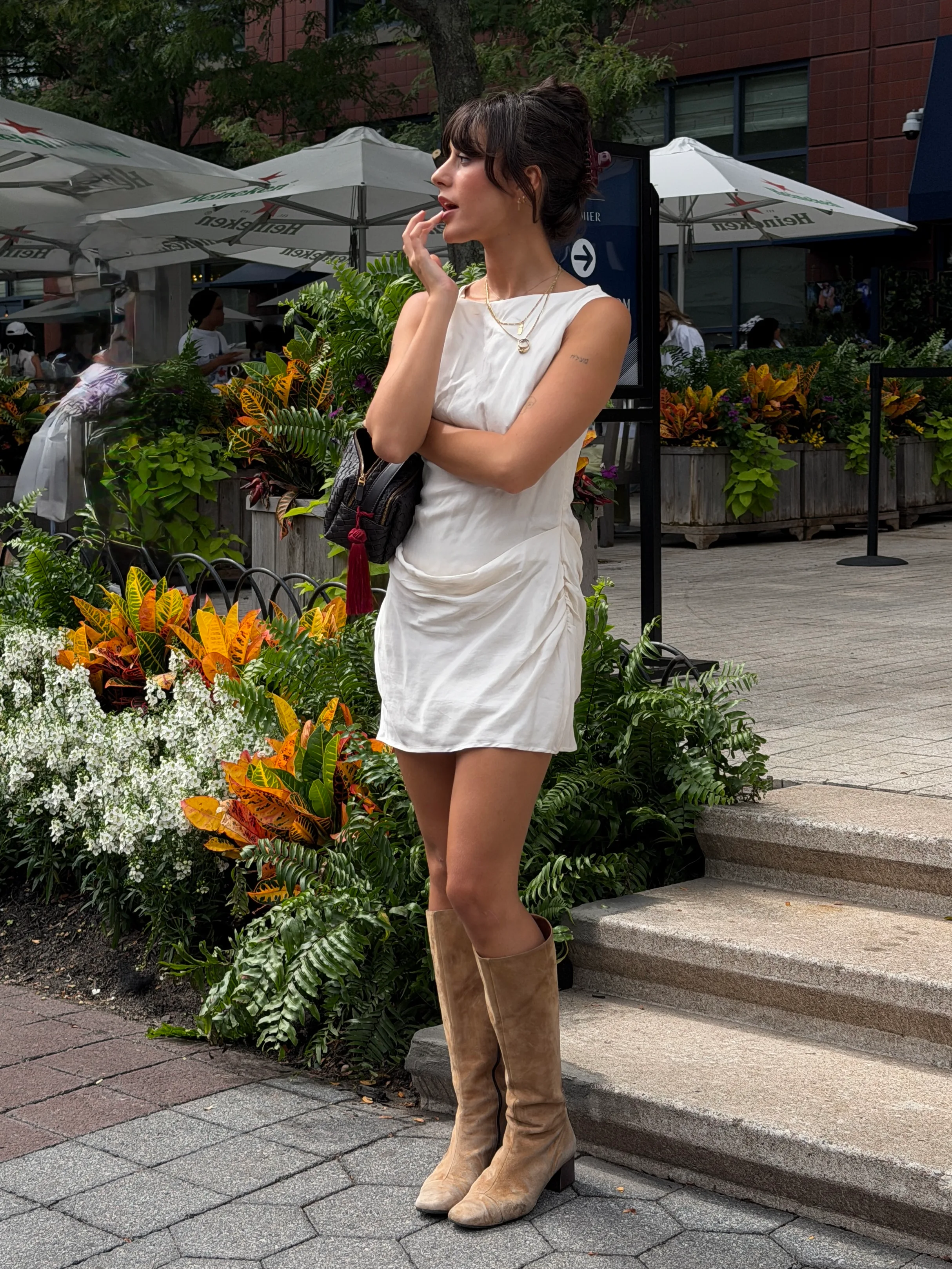 White sleeveless dress with ruched details, brown knee-high boots, and a black woven bag.