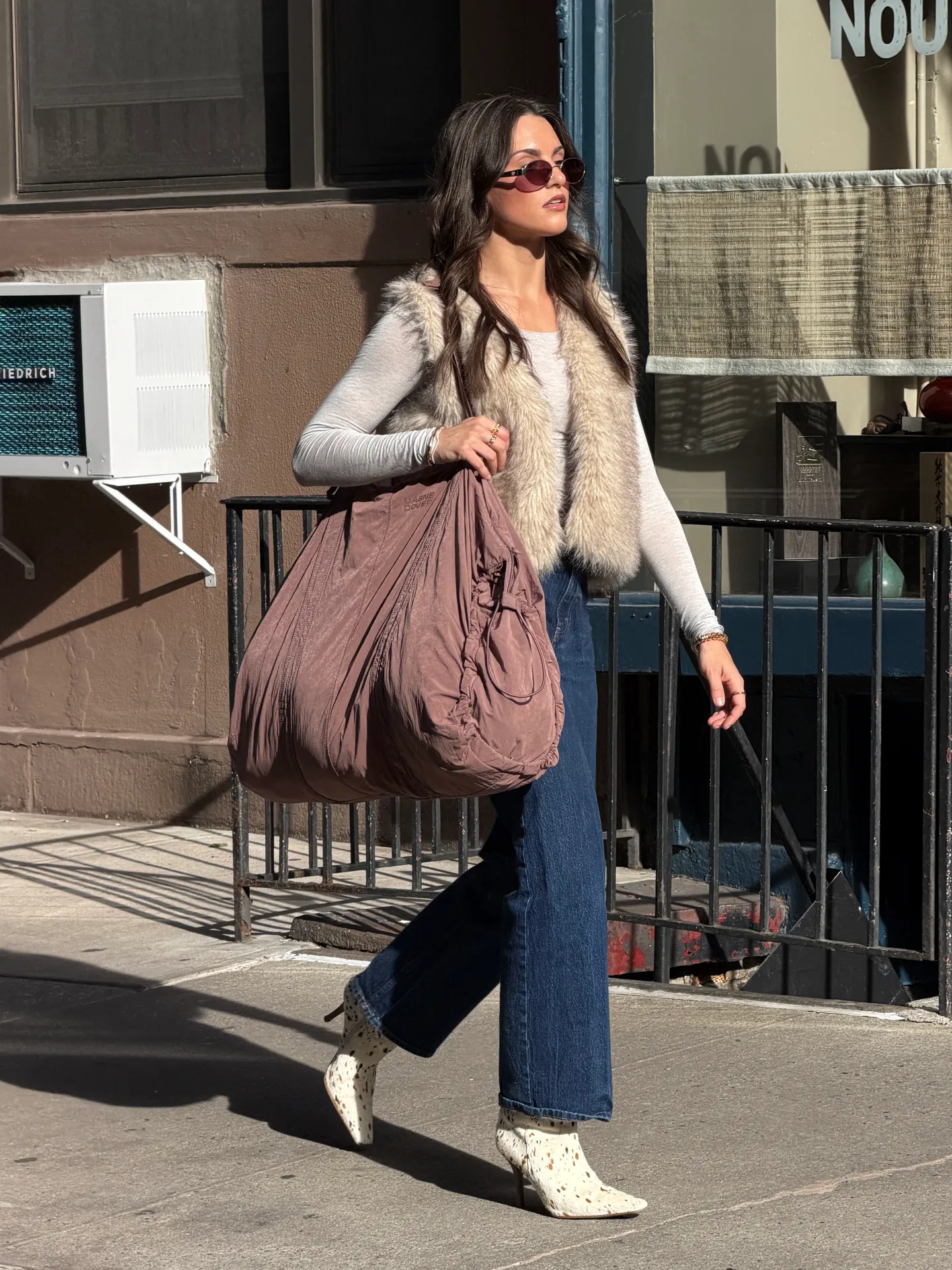 Beige faux fur vest, light gray top, wide-leg jeans, patterned boots, and pink tote bag.