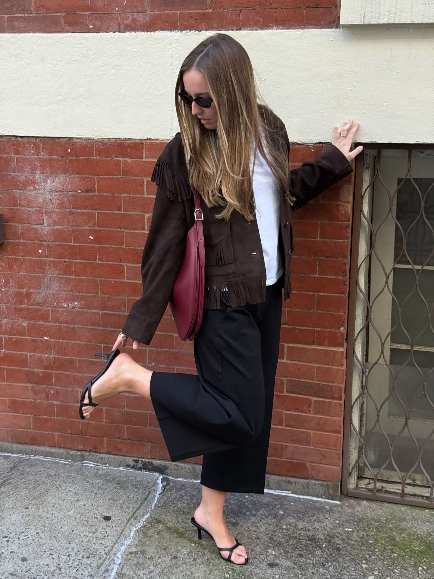 Brown fringe jacket, white top, black cropped pants, black kitten heels, and a burgundy bag.