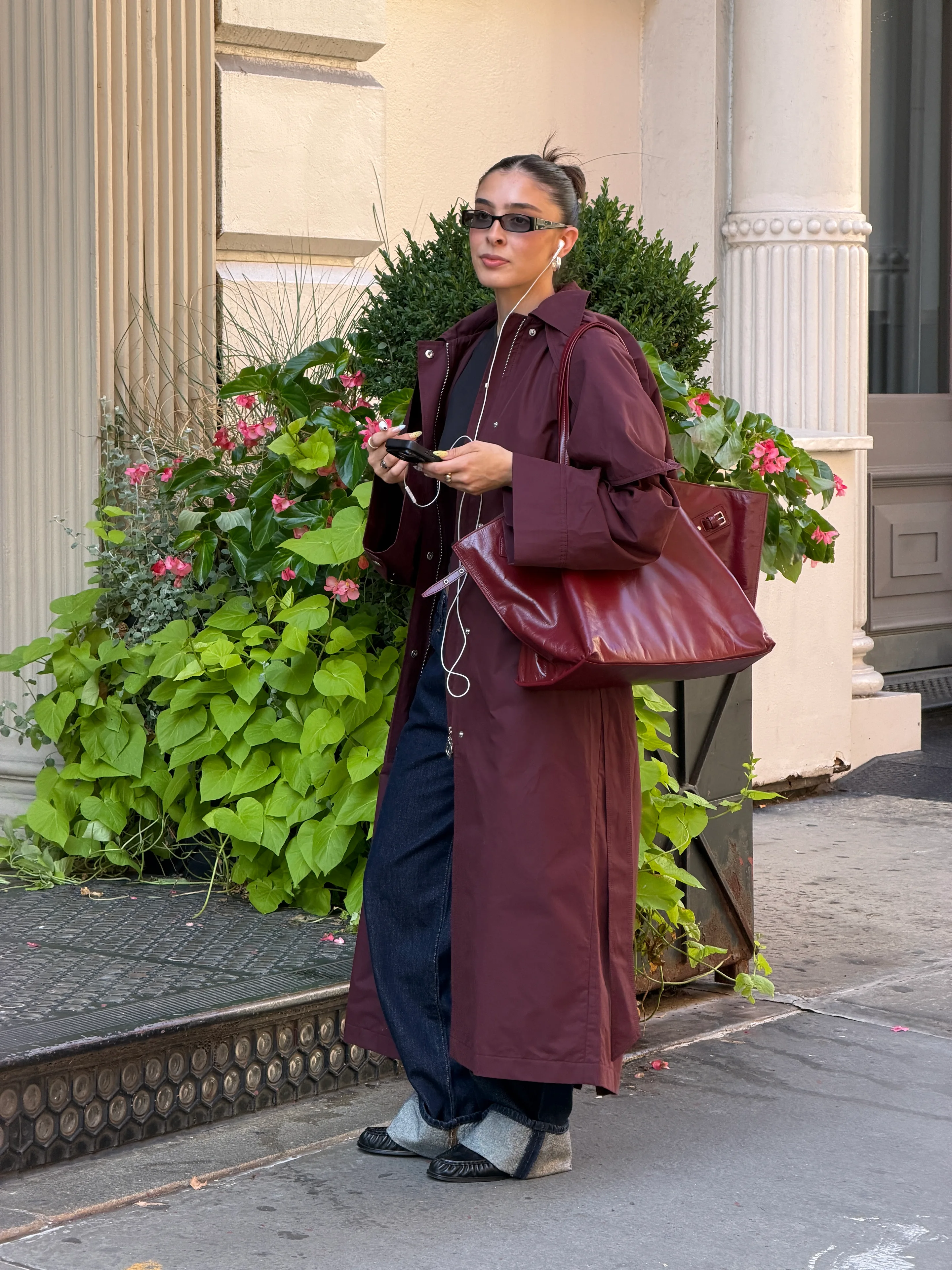 Burgundy trench coat, wide-leg jeans, black moccasins, and narrow sunglasses on a city street.