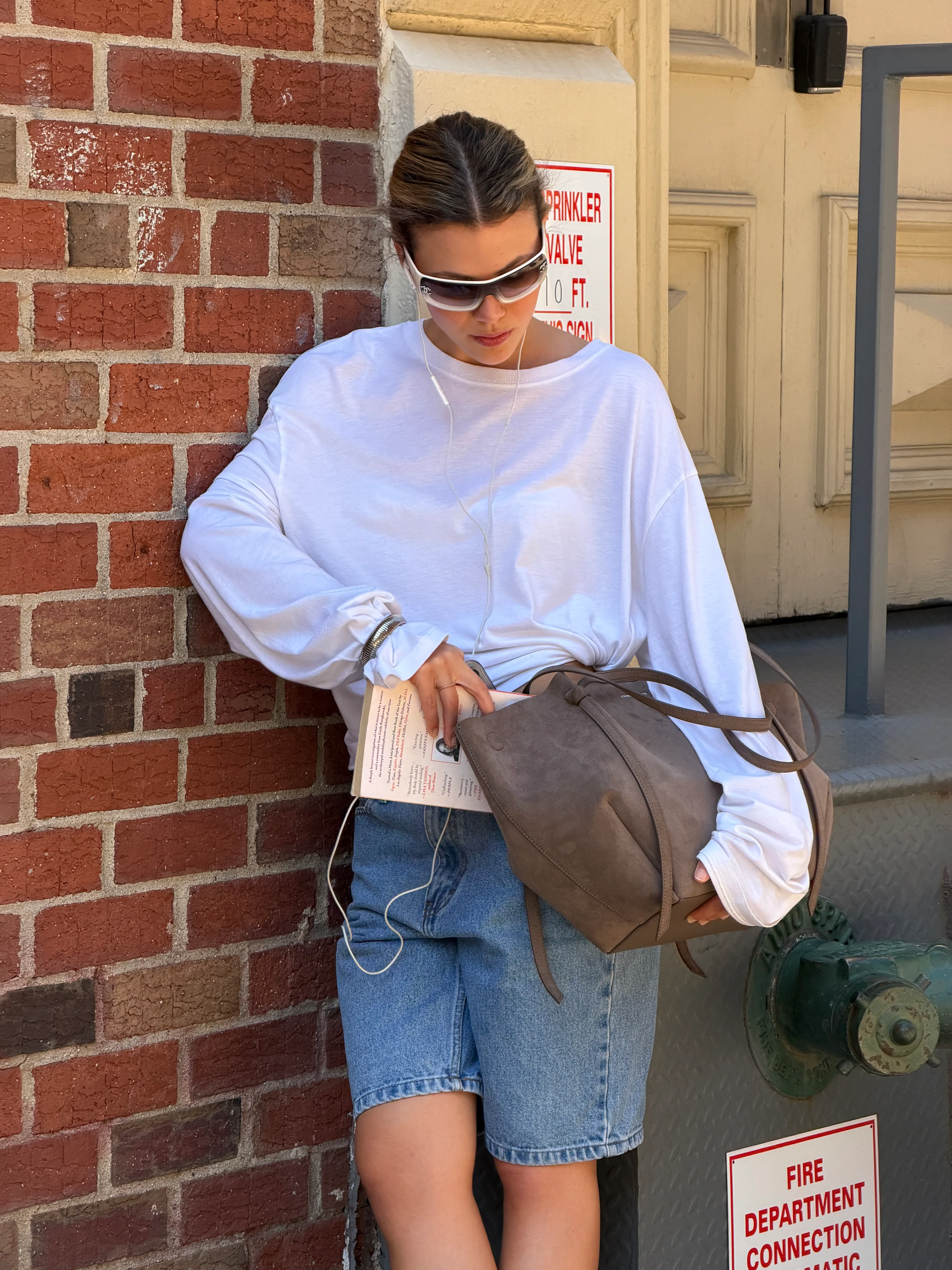 White oversized long-sleeve tee, denim shorts, gray suede tote, and futuristic sunglasses.