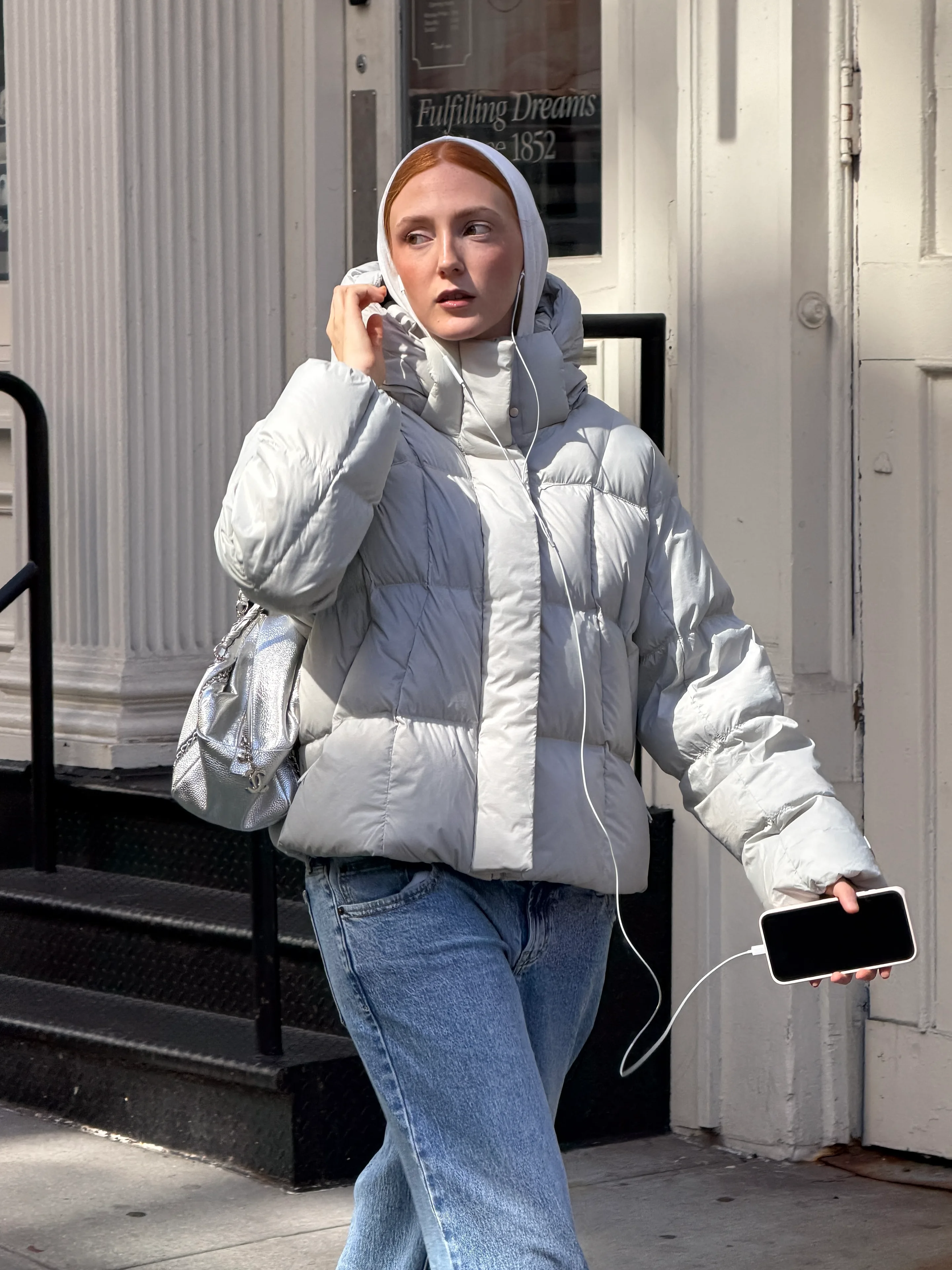 Light grey puffer jacket, white hoodie, blue jeans, and silver handbag on a street.