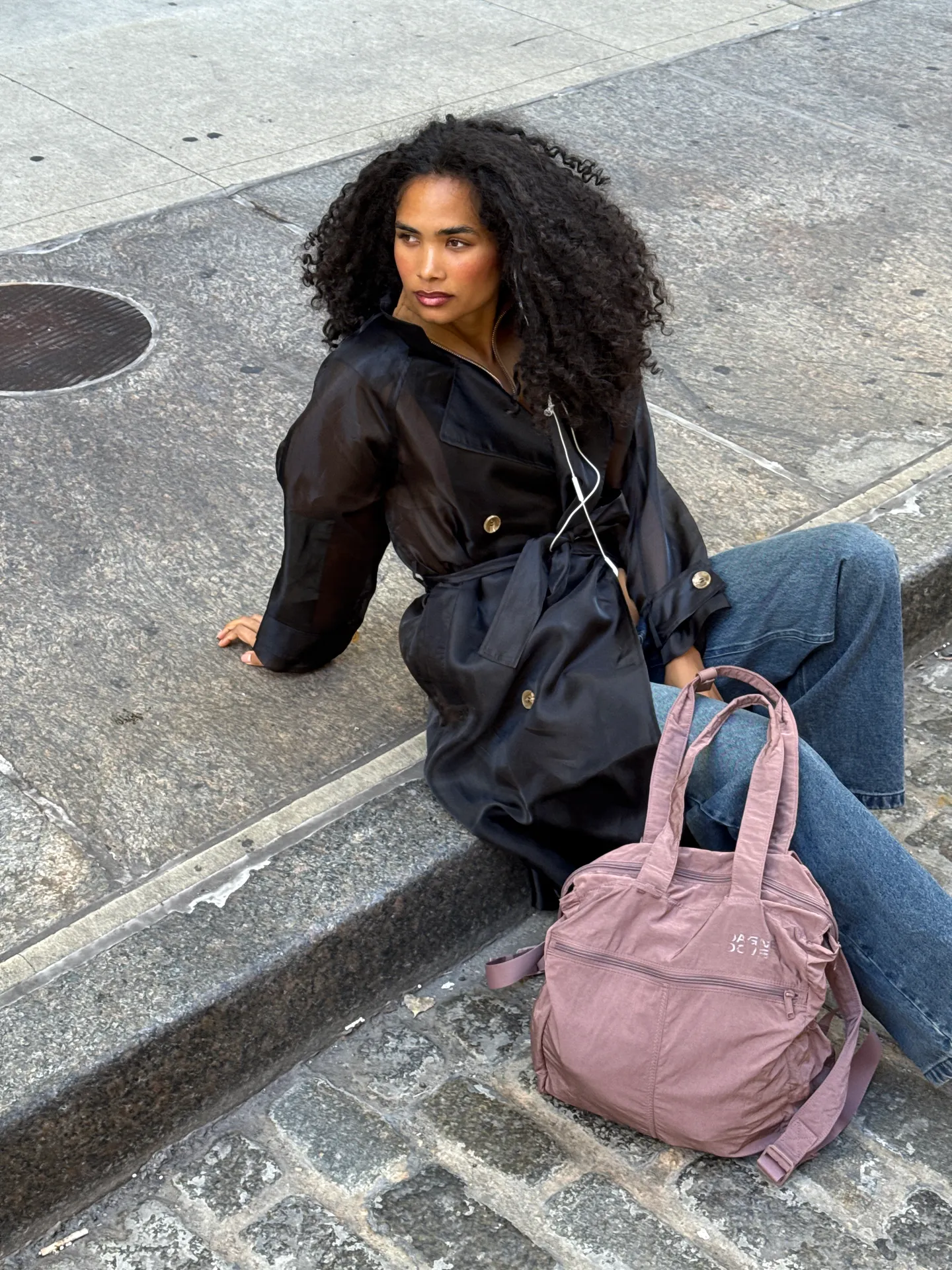 Black sheer trench coat with gold buttons, wide-leg jeans, and a mauve tote bag.