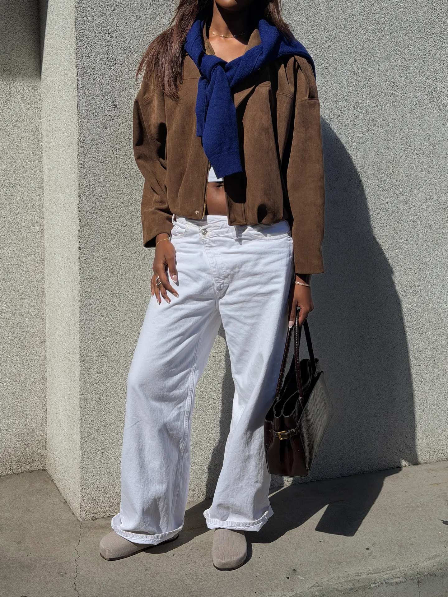 Brown suede bomber jacket, blue sweater, white wide-leg jeans, brown bag, and taupe clogs.