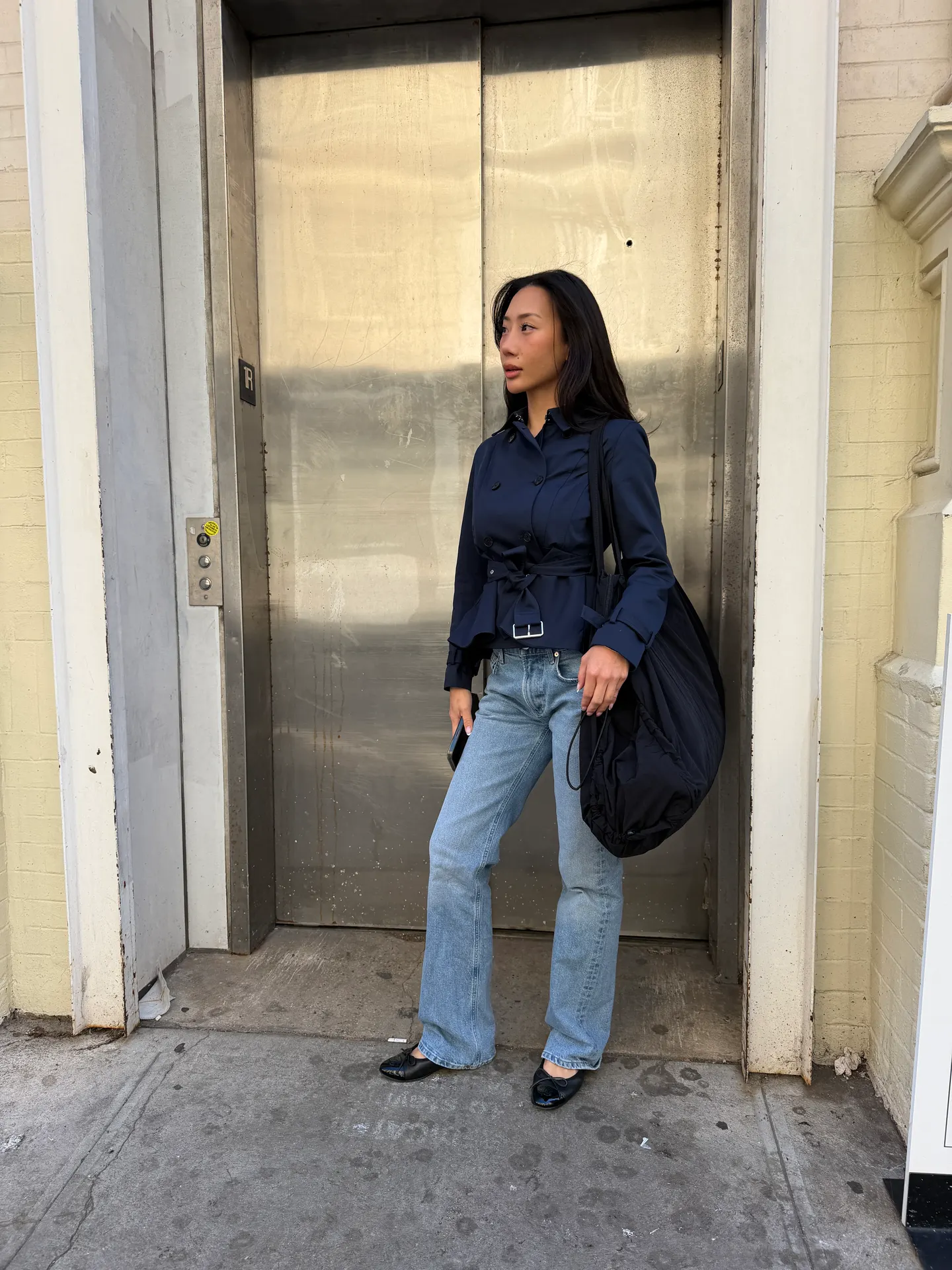 Navy belted jacket with light wash jeans, black ballet flats, and large black tote bag.
