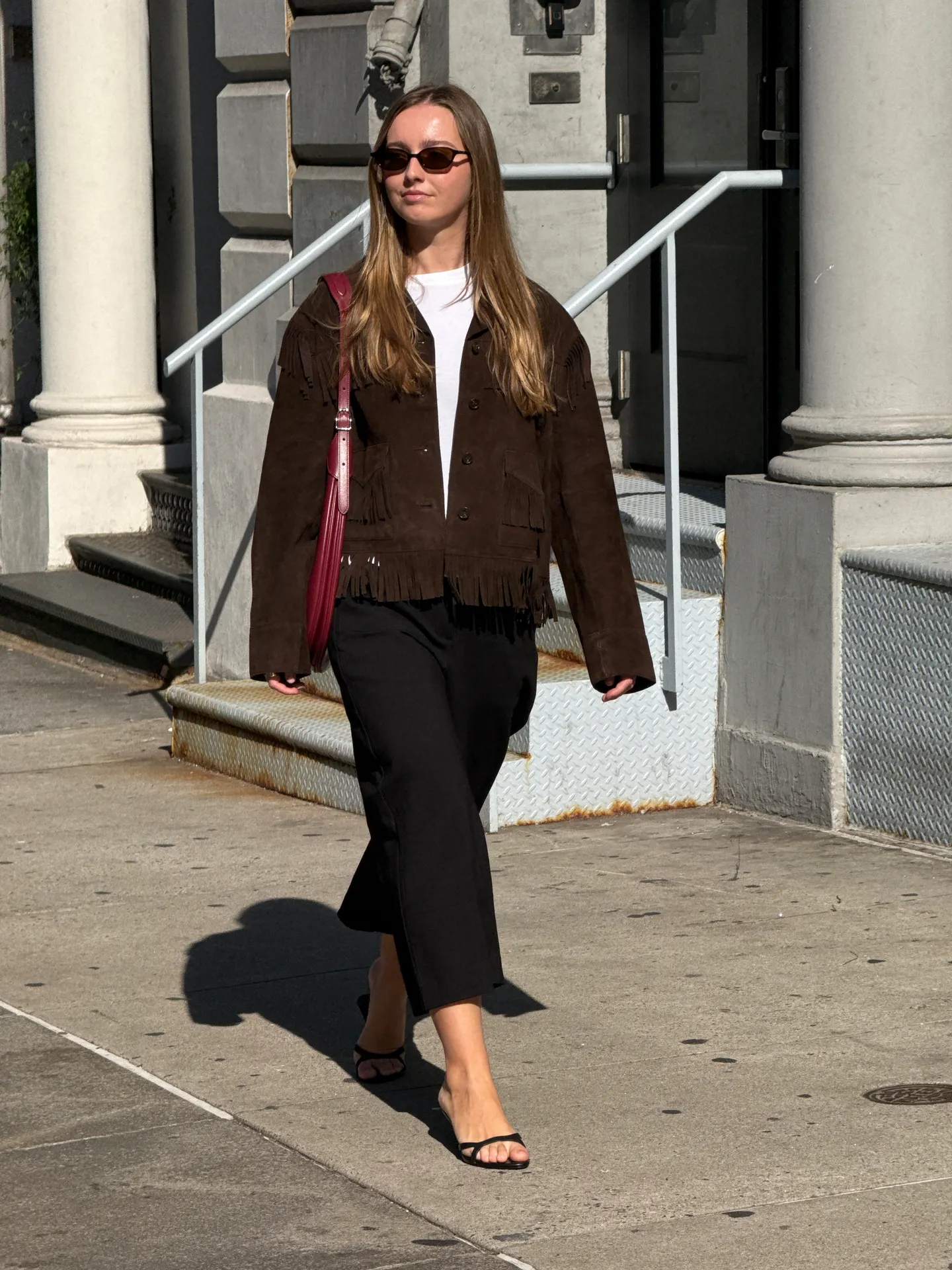 Brown suede fringe jacket, white tee, black pants, black sandals, and red shoulder bag.