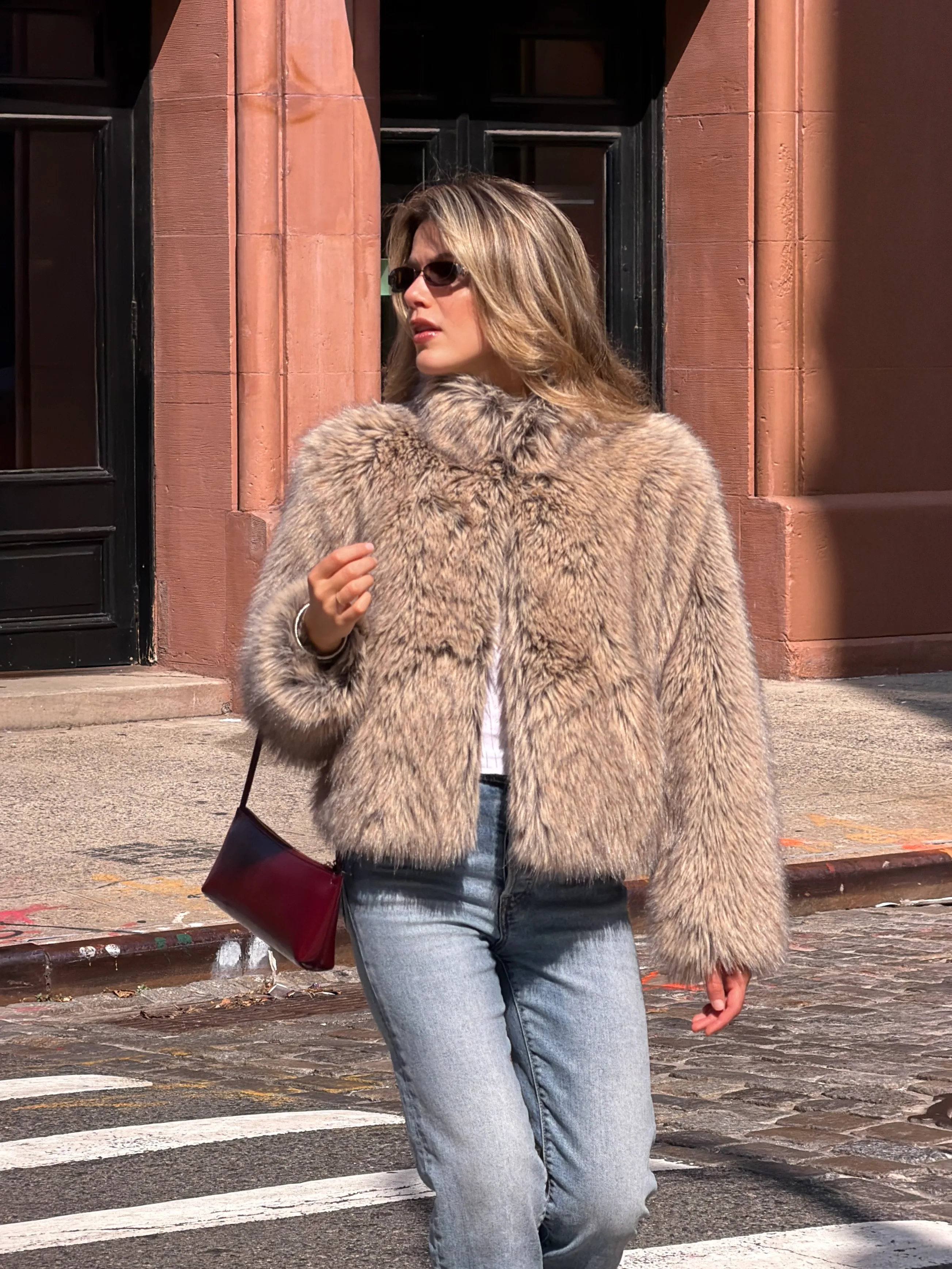 Brown faux fur jacket with light wash jeans and a burgundy bag on a city street.