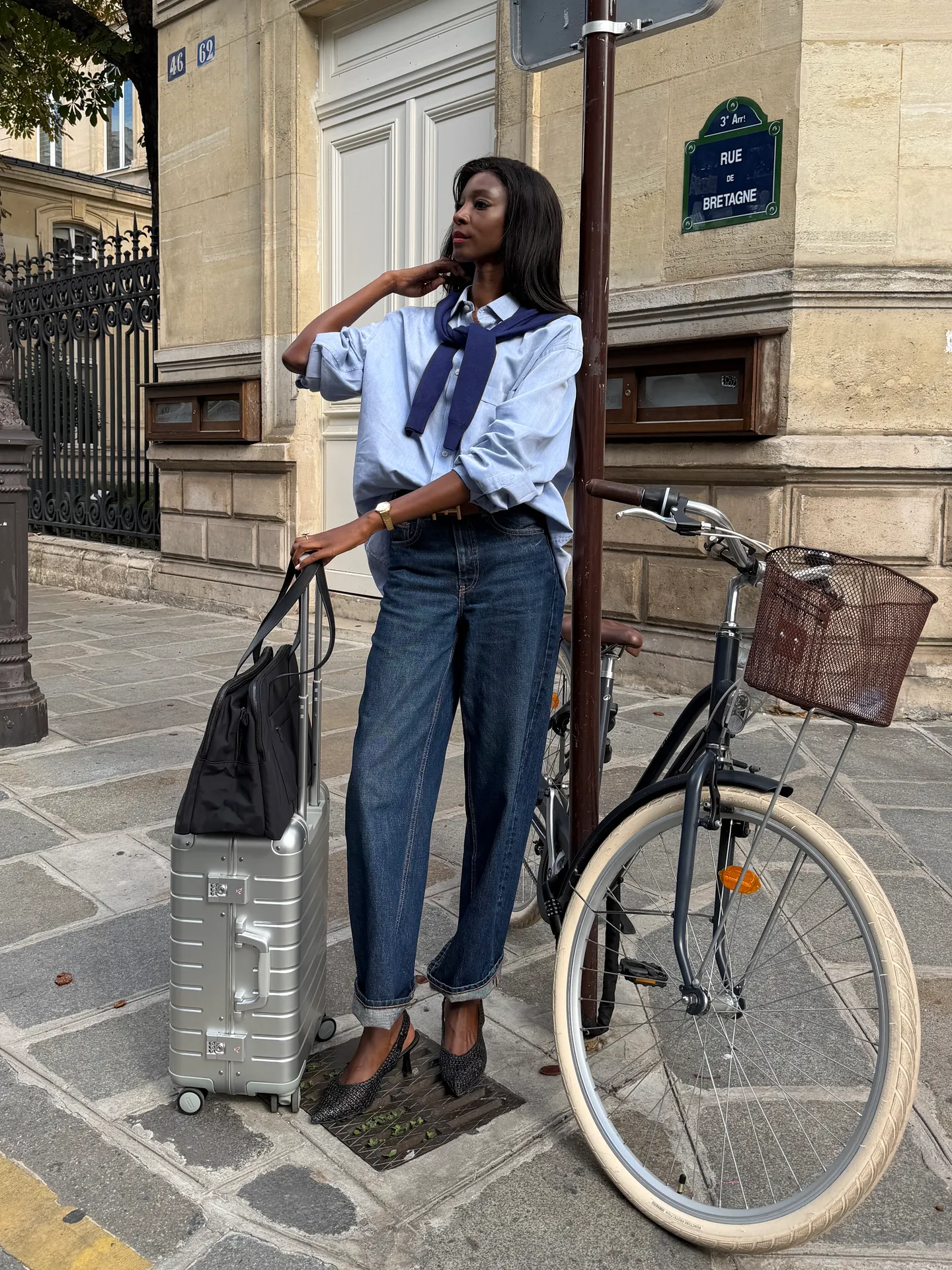 Blue button-down shirt, dark wash jeans, black slingback heels, silver suitcase, and a bike.