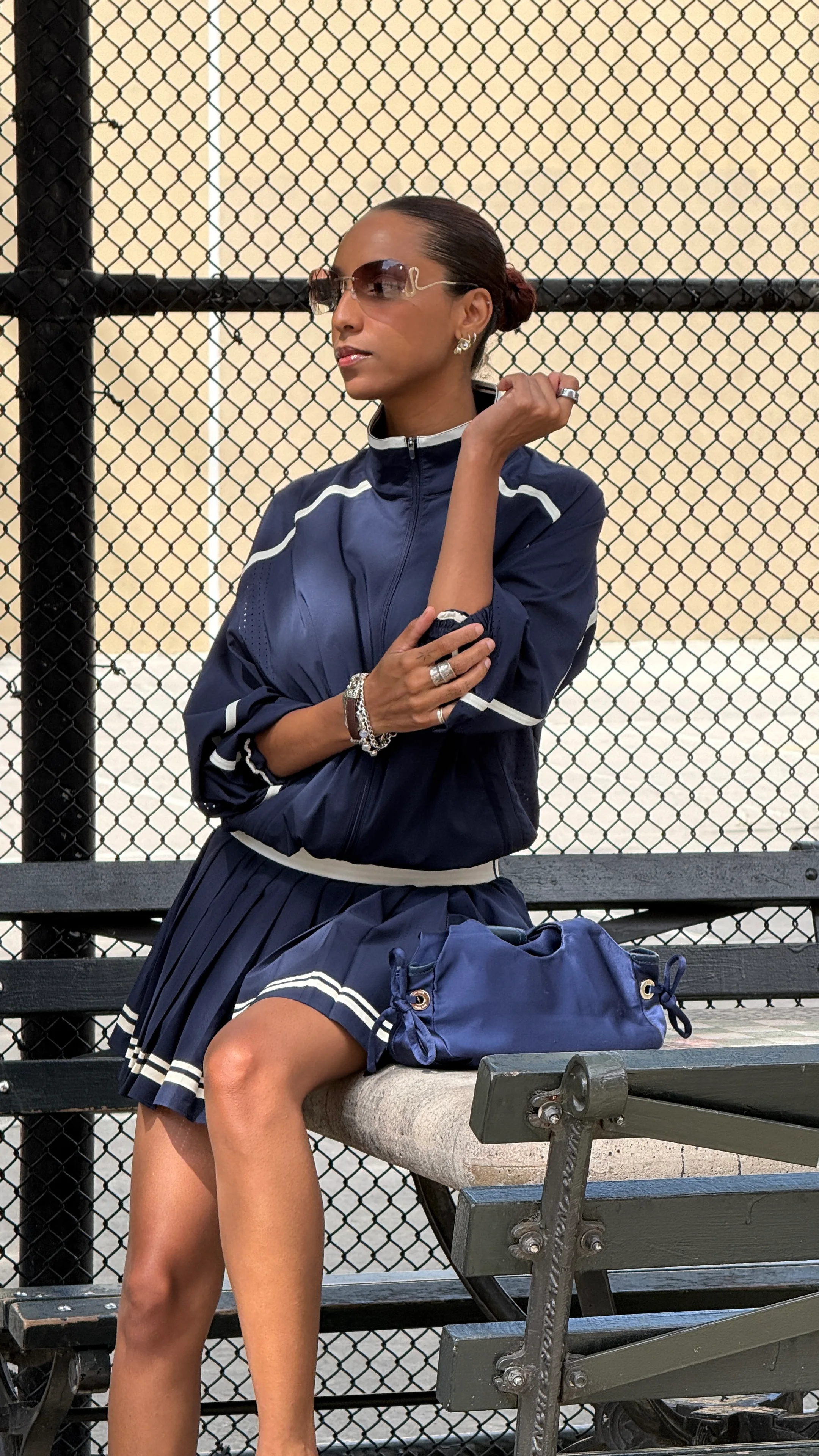 Navy blue tracksuit with white stripes, pleated skirt, sunglasses, and a matching bag.