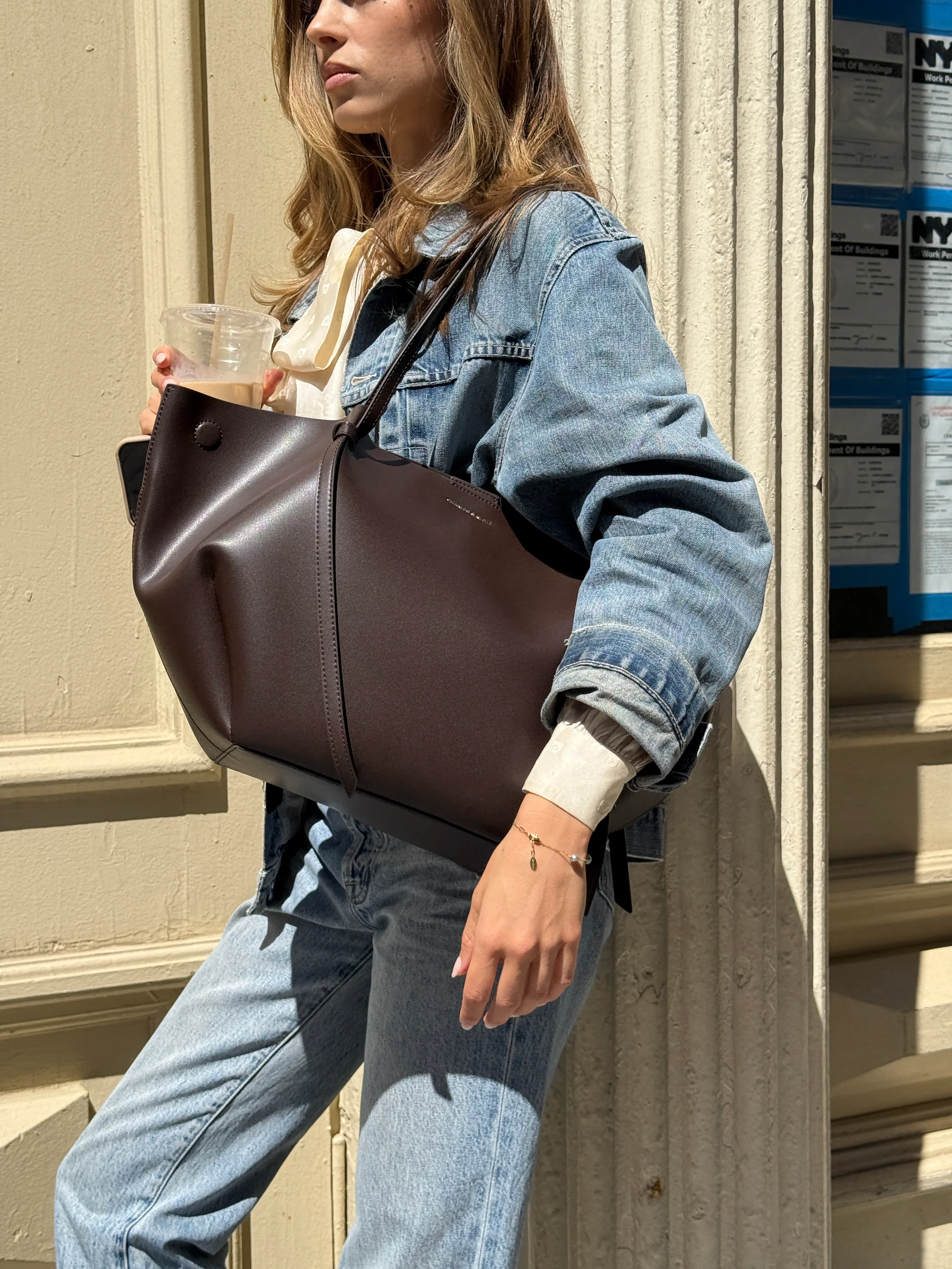 Light wash denim jacket and jeans, brown tote bag, pearl bracelet, holding coffee.