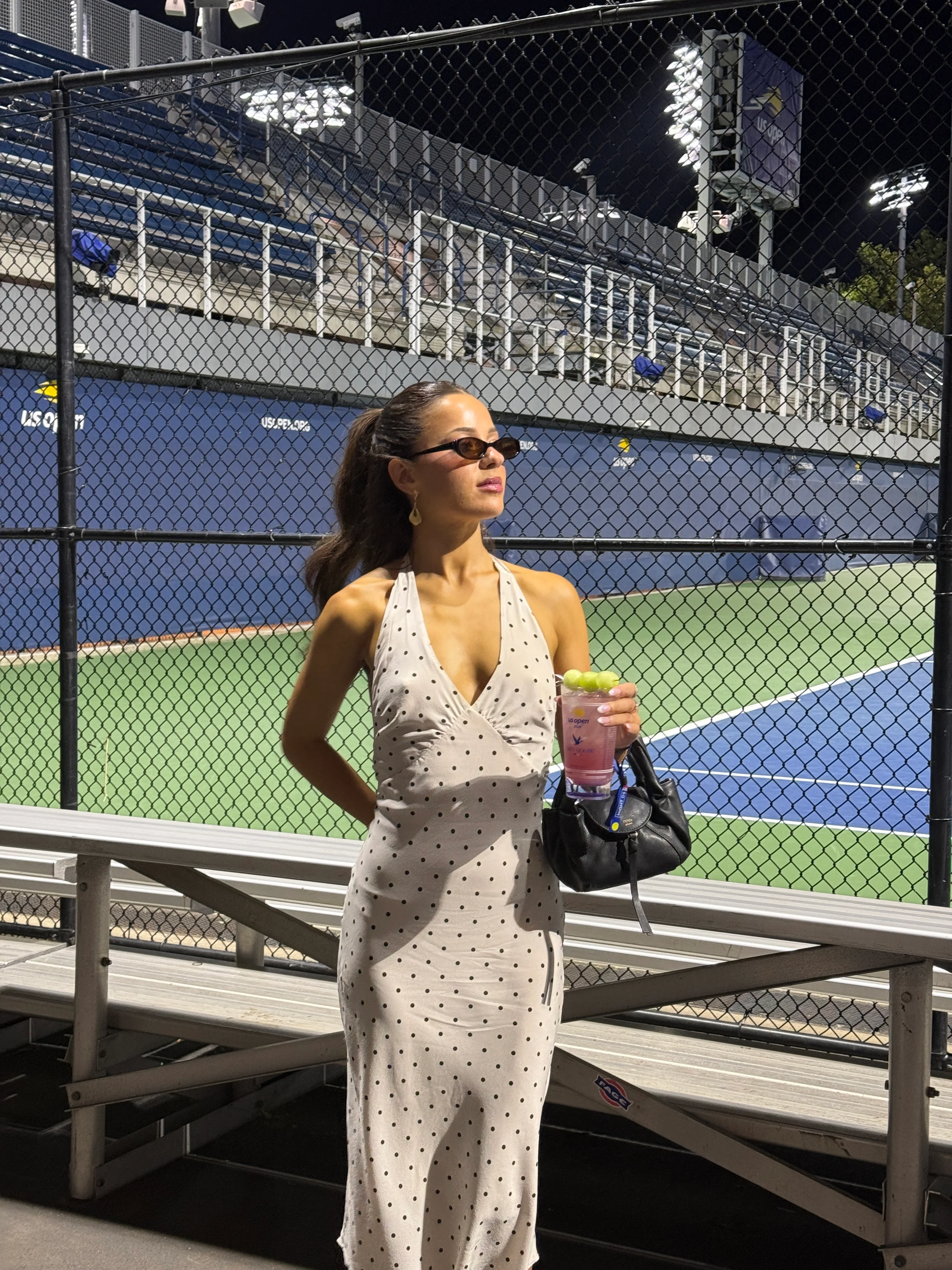 White polka dot halter dress, dark sunglasses, black handbag, holding a drink at a tennis court.