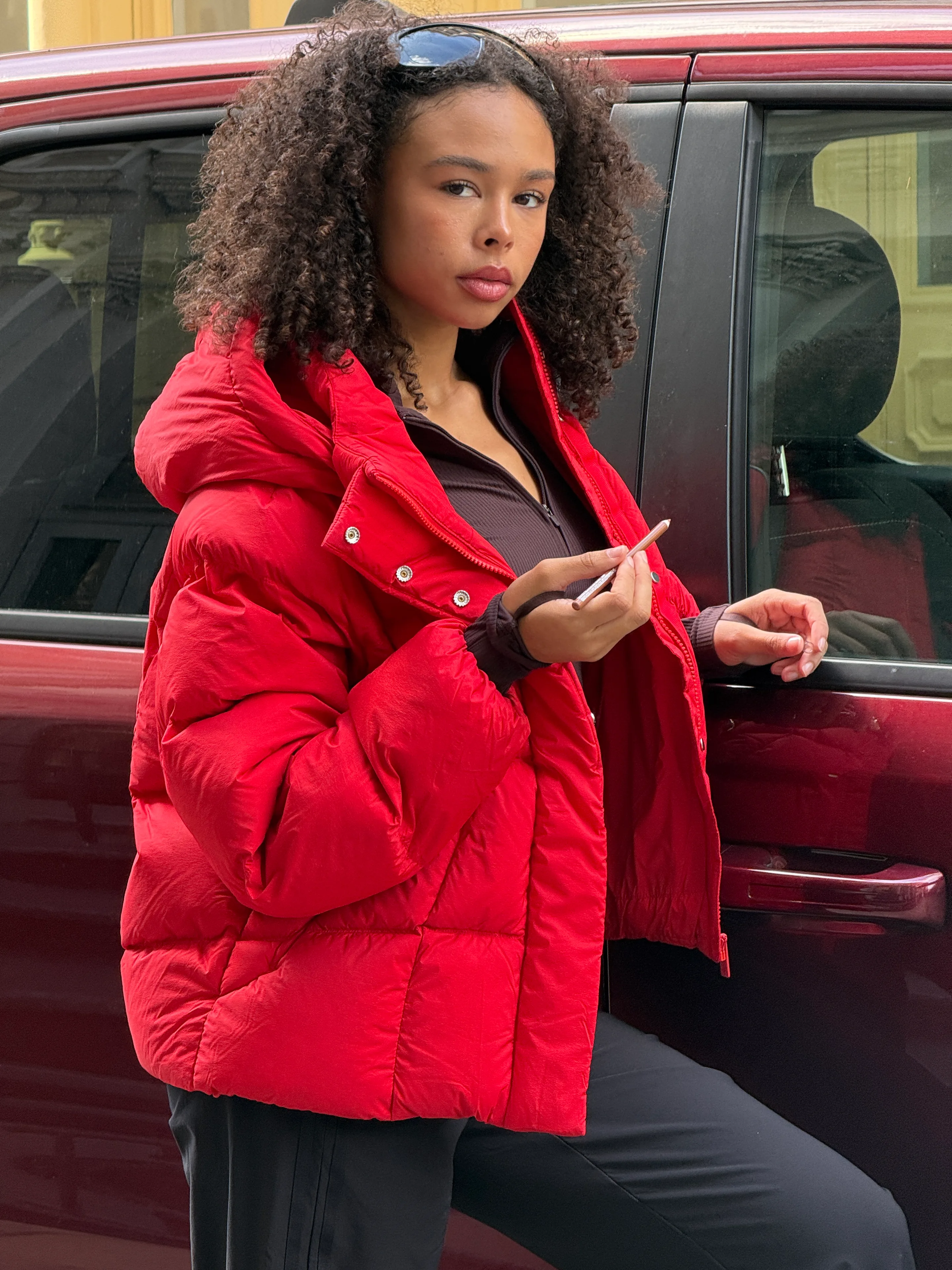 Red puffer jacket over brown top and black pants, sunglasses on head.