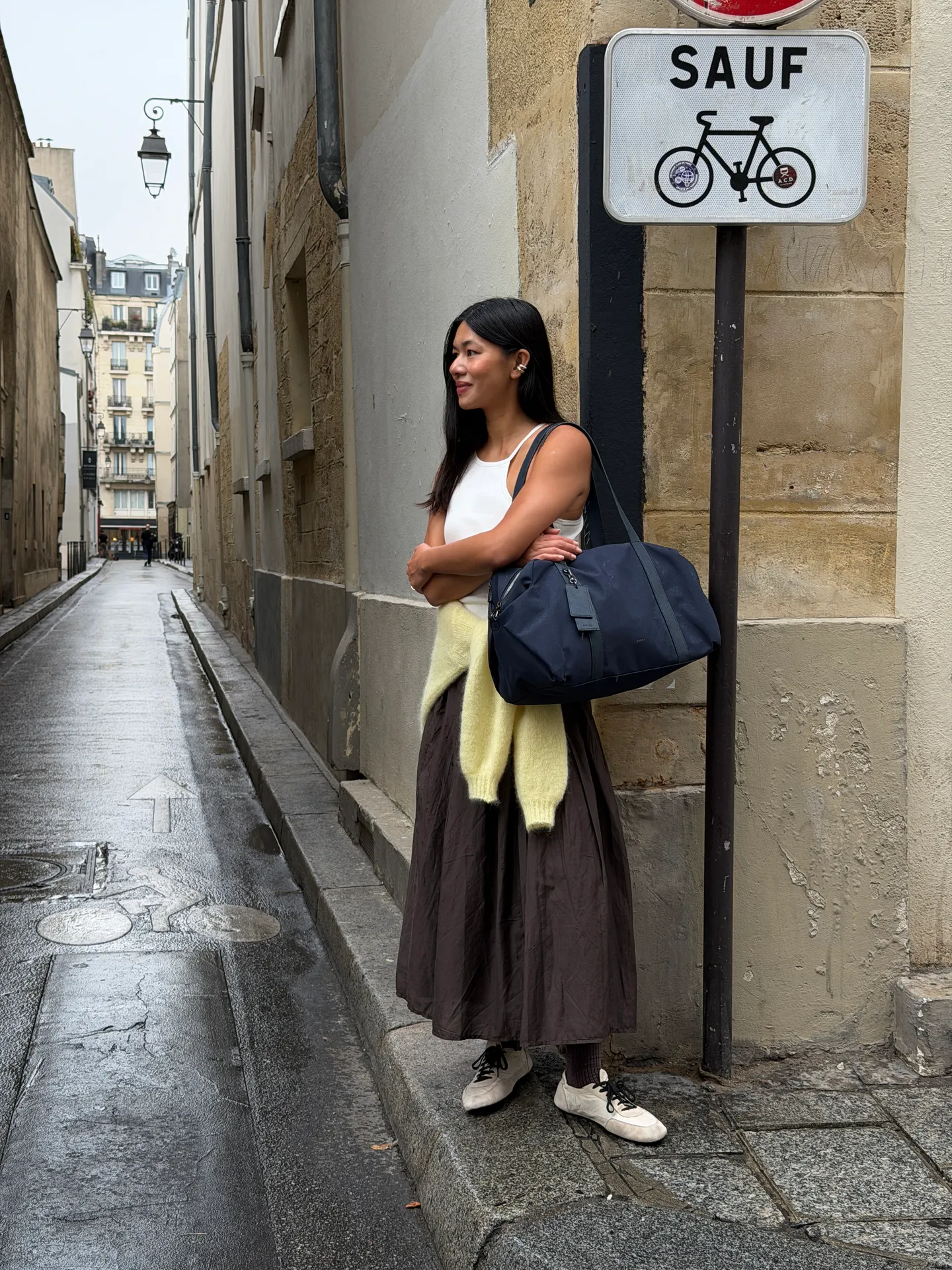 White tank top, brown maxi skirt, yellow sweater, navy duffel bag on a wet street.
