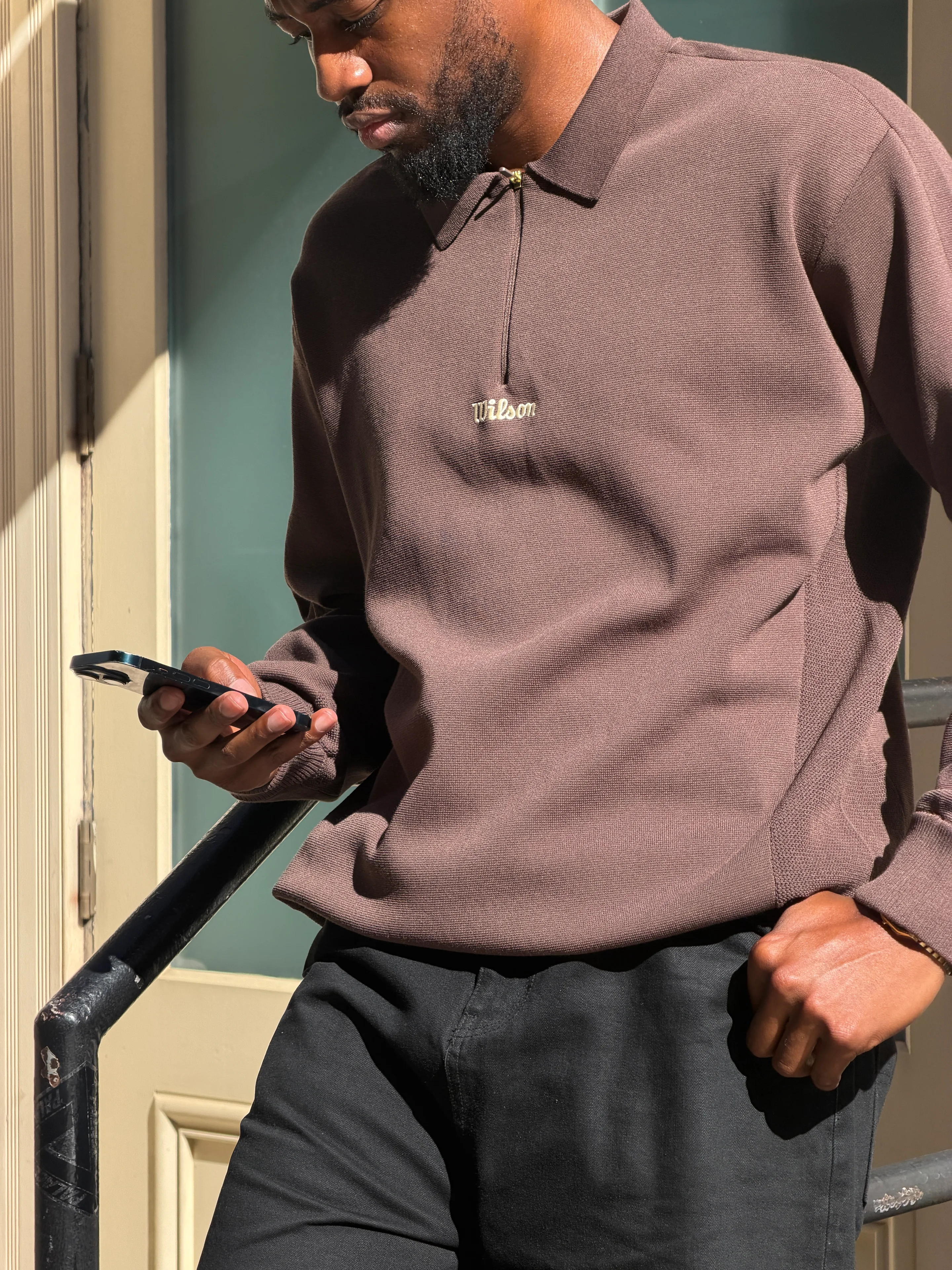 Brown Wilson zip-up polo shirt and black trousers, holding a smartphone.