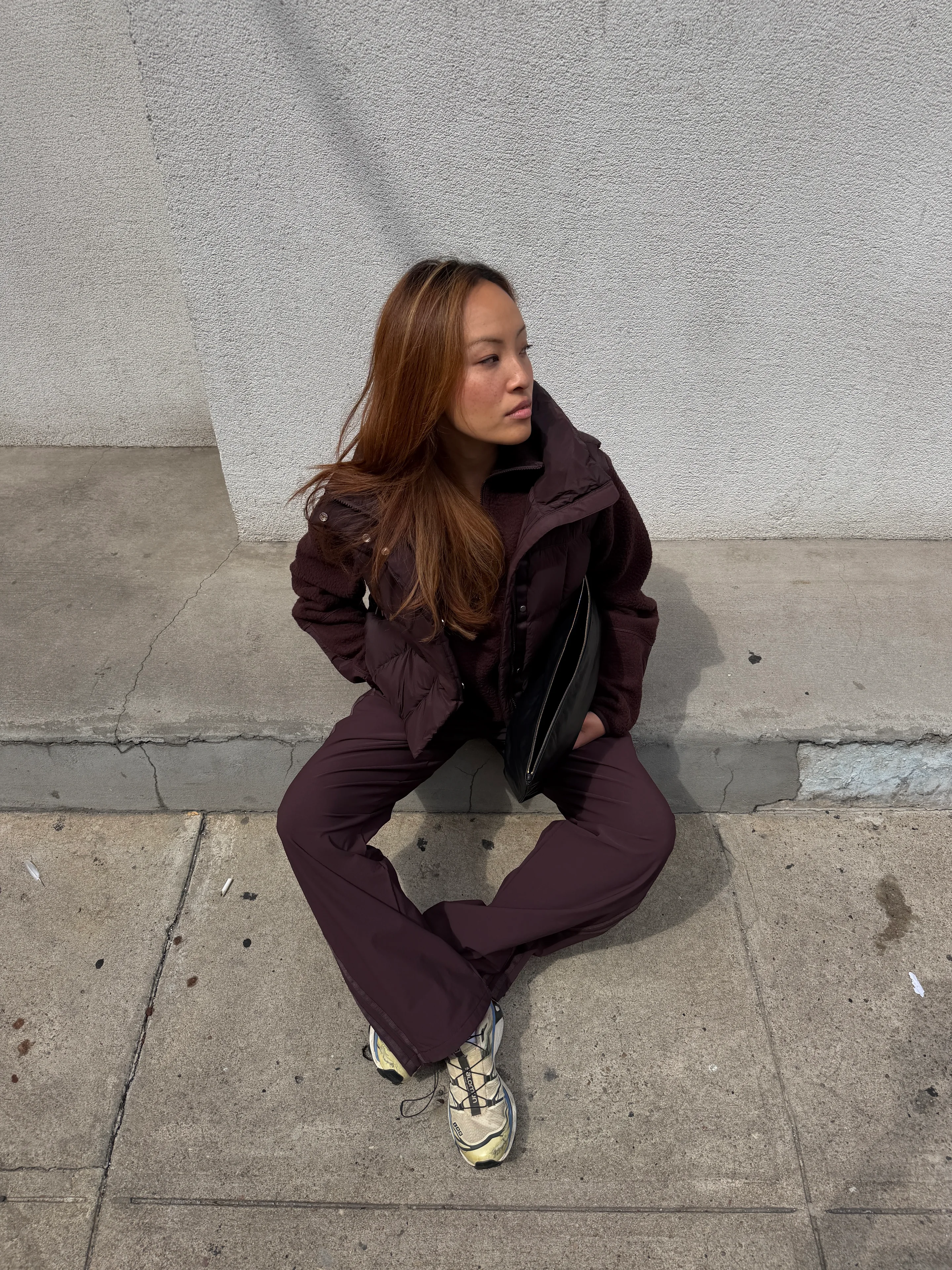 Brown puffer vest, fleece top, and pants with light sneakers, holding a black clutch.