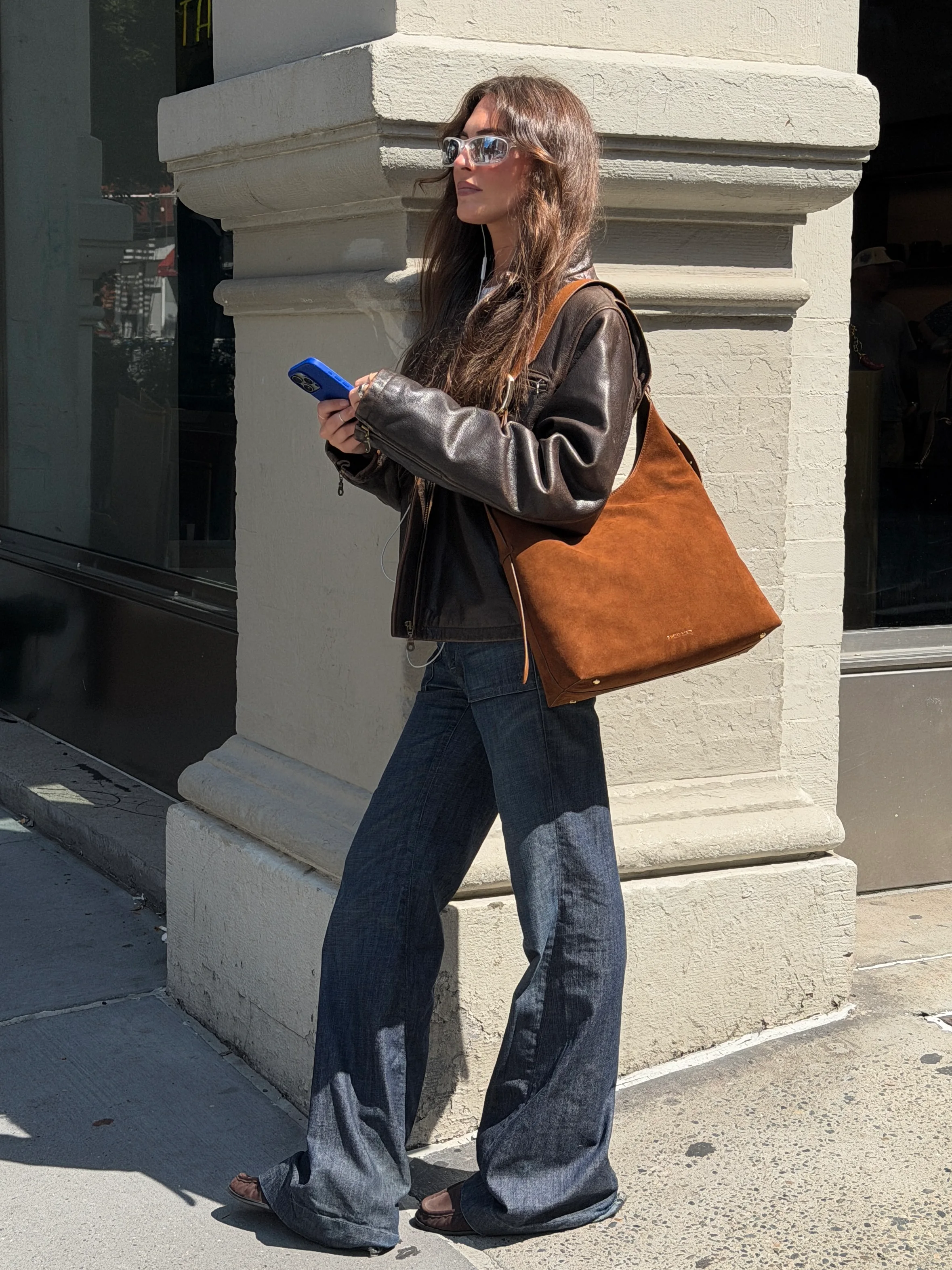 Brown leather jacket, wide-leg jeans, brown suede bag, and slim sunglasses on a sunny street.