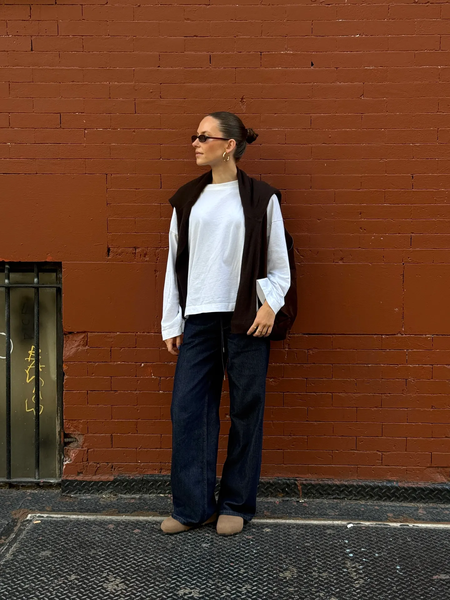 White long-sleeve top, dark brown sweater, wide-leg jeans, and tan loafers against brick wall.